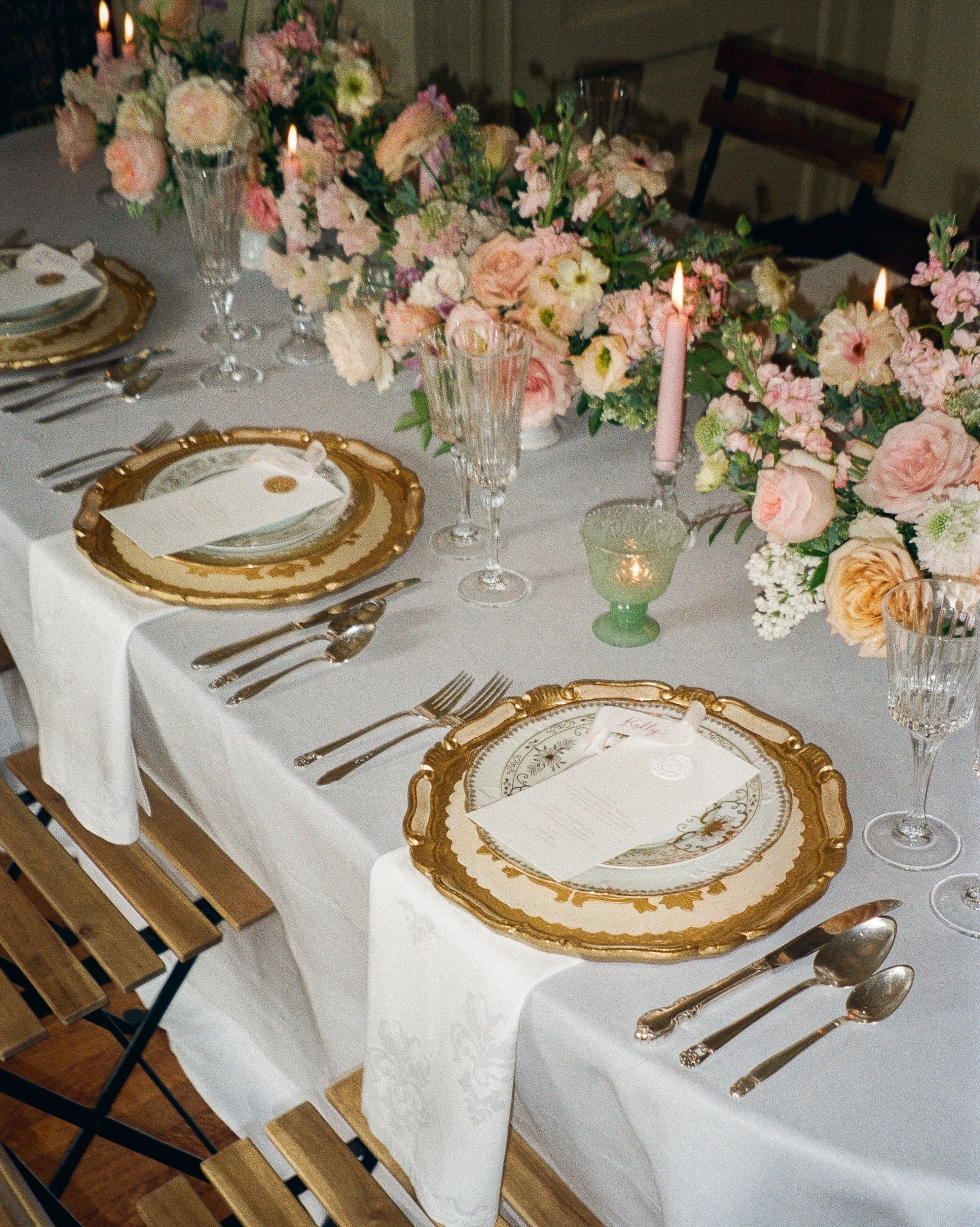 A long rectangular table covered in a white table cloth with elaborate gold place settings and wooden chairs. Pink taper candles and green translucent glass votives are lit on the table. 3 flower centerpieces in pink a decorate the table.