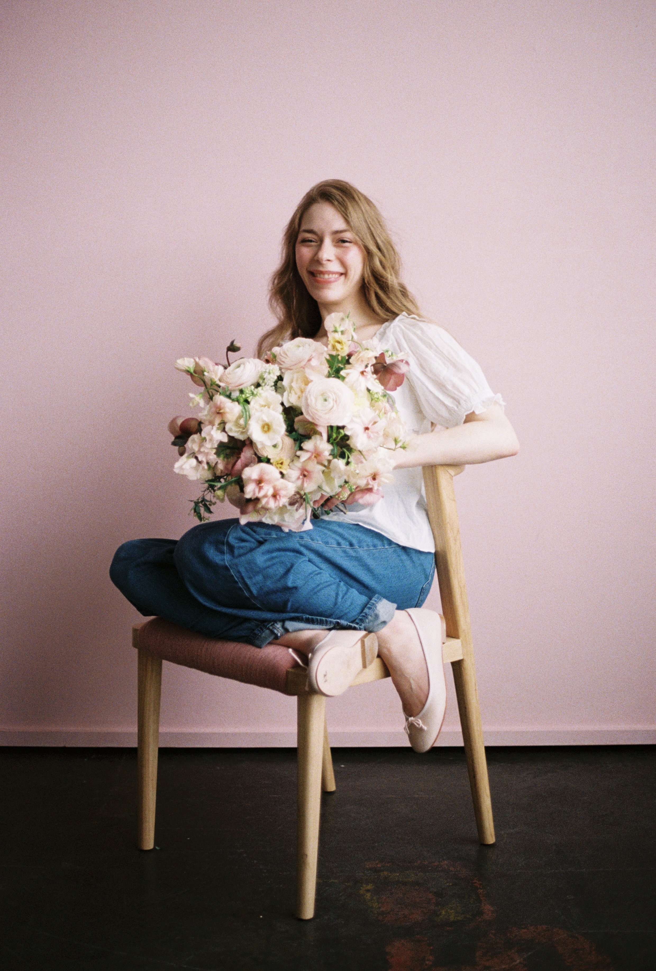 A girl wearing a white shirt, blue jeans, and light pink ballet flats sits on a pink stool holding a large garden-style bouquet. The bouquet is pink and mauve. The background is light rose pink.