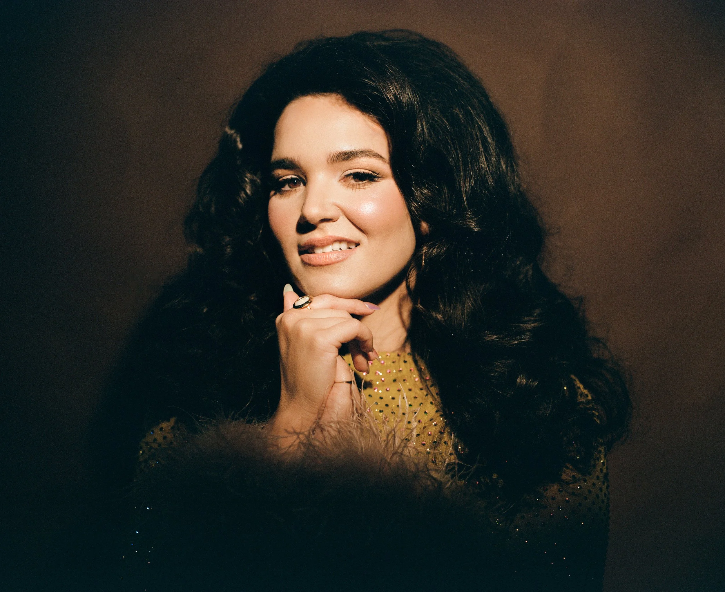 Portrait of a woman with long, dark, curly hair, winking, and smiling slightly, wearing a gold dress with small sparkles, and a ring on her finger, against a dark background.