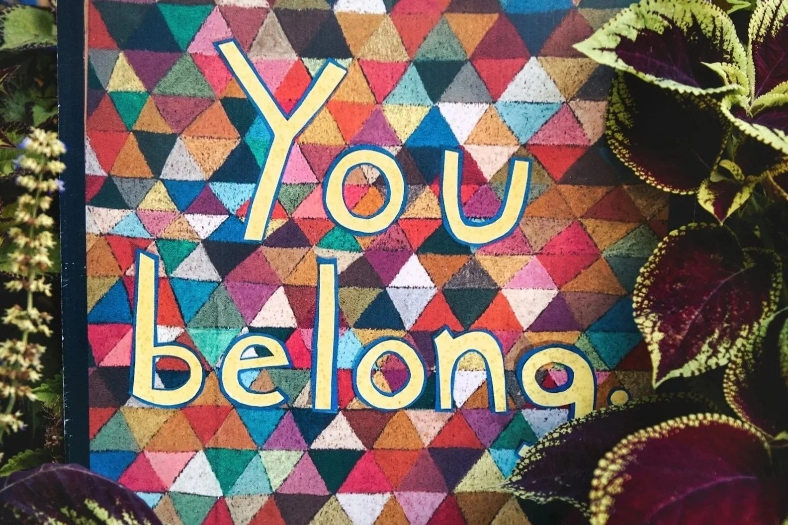 Colorful mosaic background with the phrase "You belong" written in large, yellow letters outlined in blue, surrounded by green and purple foliage on the sides.