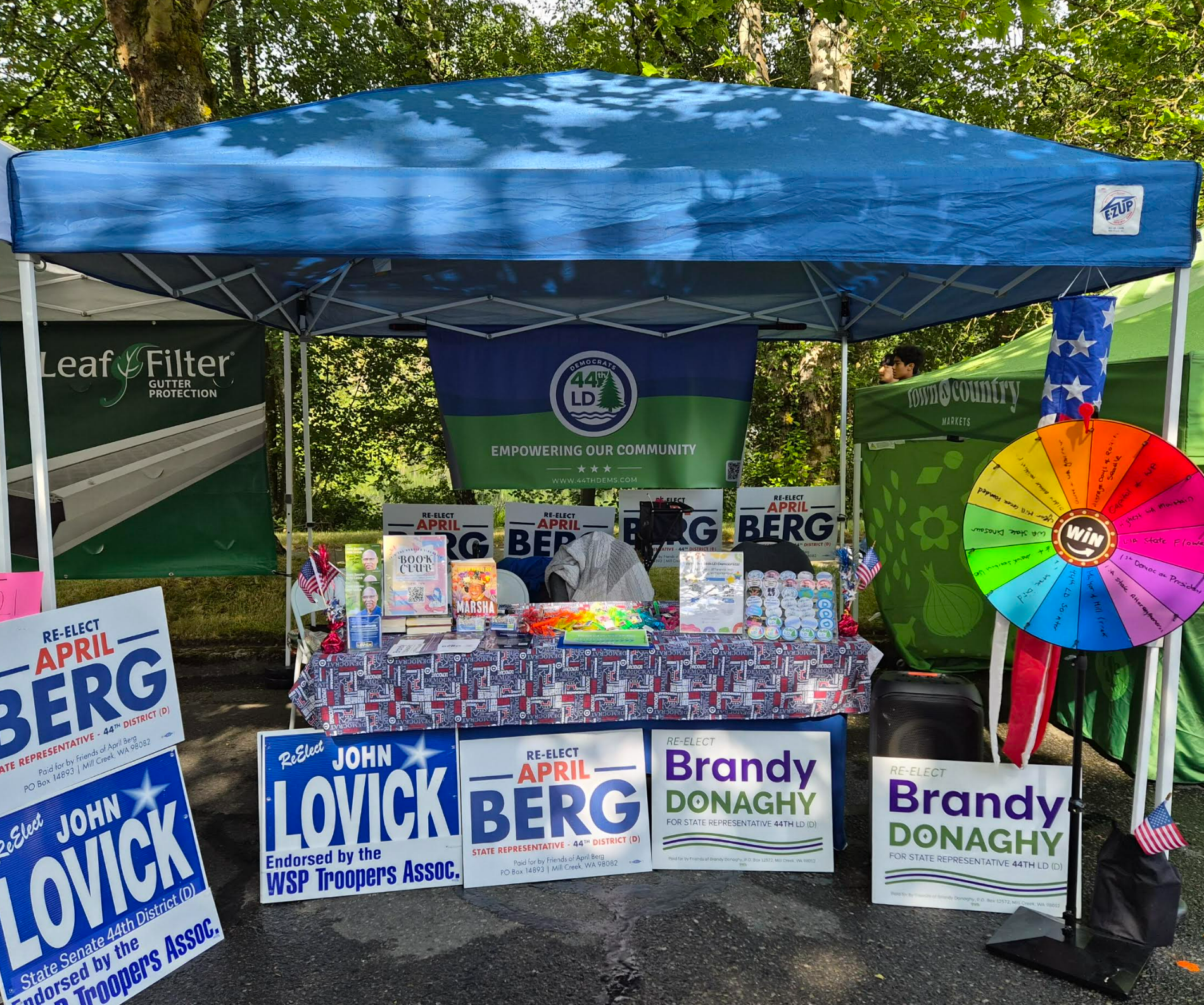 A political campaign booth with signs and banners supporting candidates John Lovick and Brandy Donaghy. There is a blue canopy, a table covered with campaign materials, a colorful spin wheel, and flags.