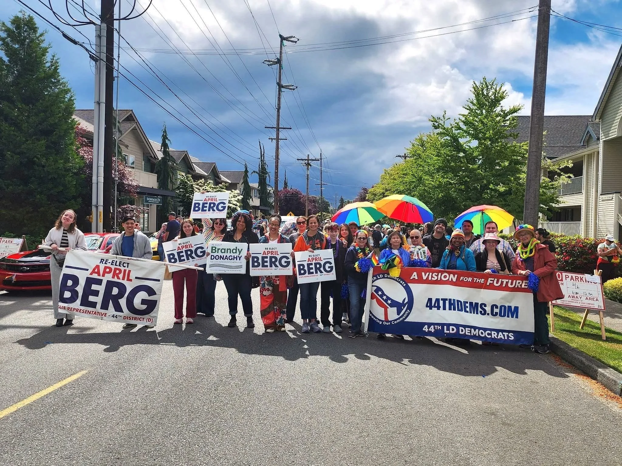 A group of people participating in a parade or rally, holding signs that support political candidates for the 44th district, with some holding rainbow-colored umbrellas, in a residential street lined with trees and houses, under a partly cloudy sky.