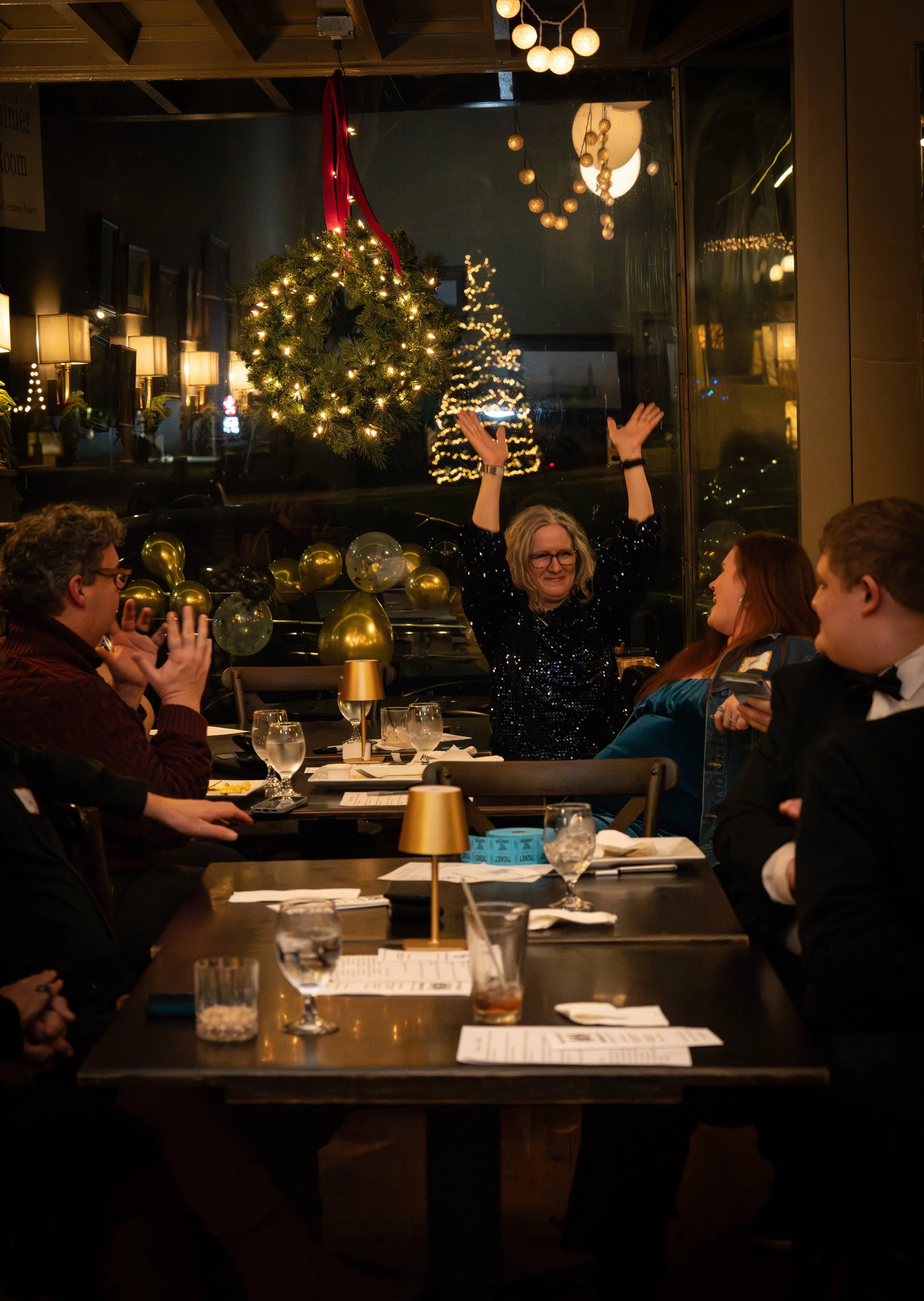 People sitting at a dinner table decorated with gold and black balloons, celebrating Christmas with a woman raising her hands, a Christmas wreath with lights, and a Christmas tree visible through the window.