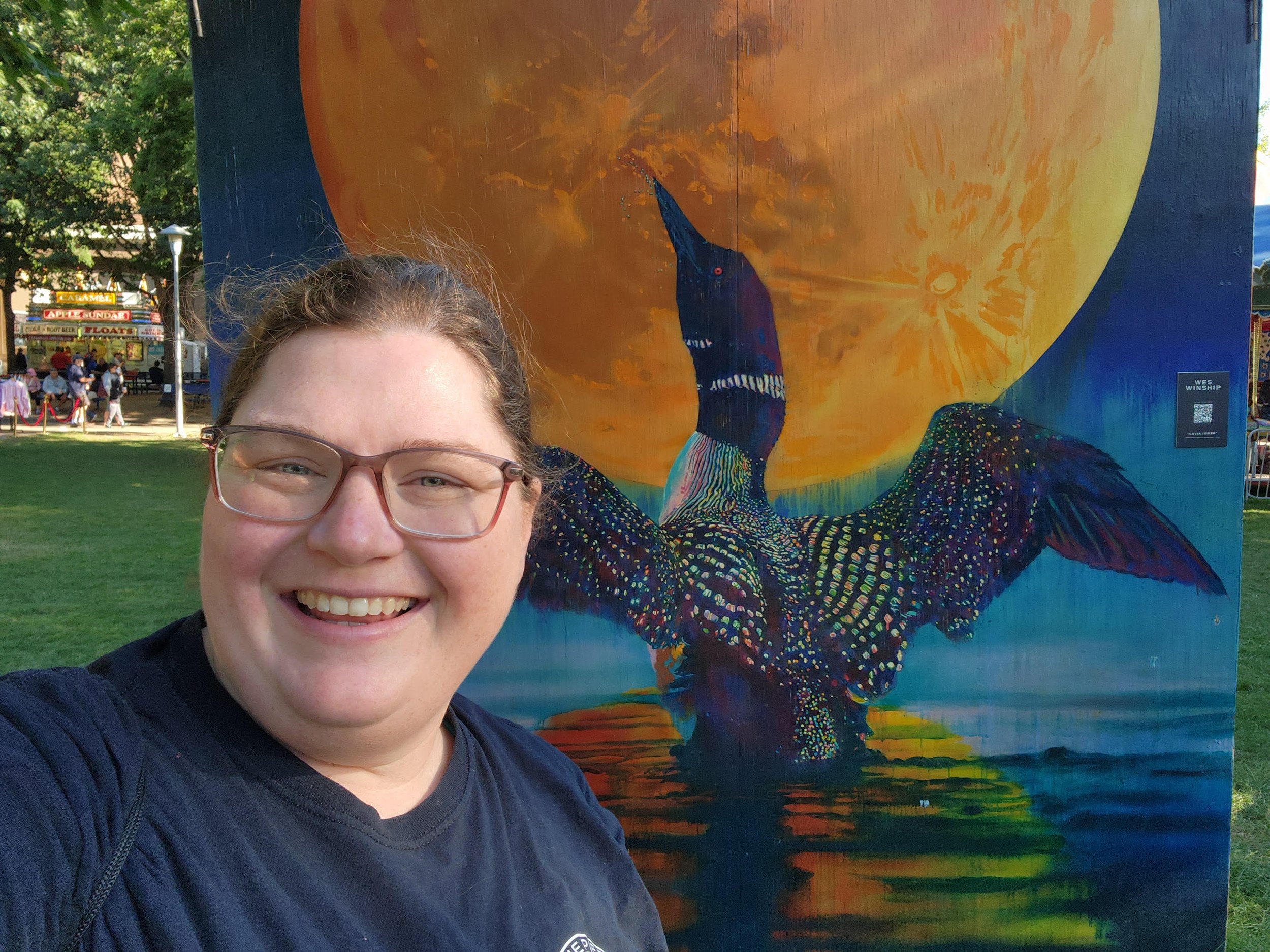 A woman with glasses smiling for a selfie in front of a colorful mural of a dragon-like creature with a large yellow moon in the background at an outdoor fair.