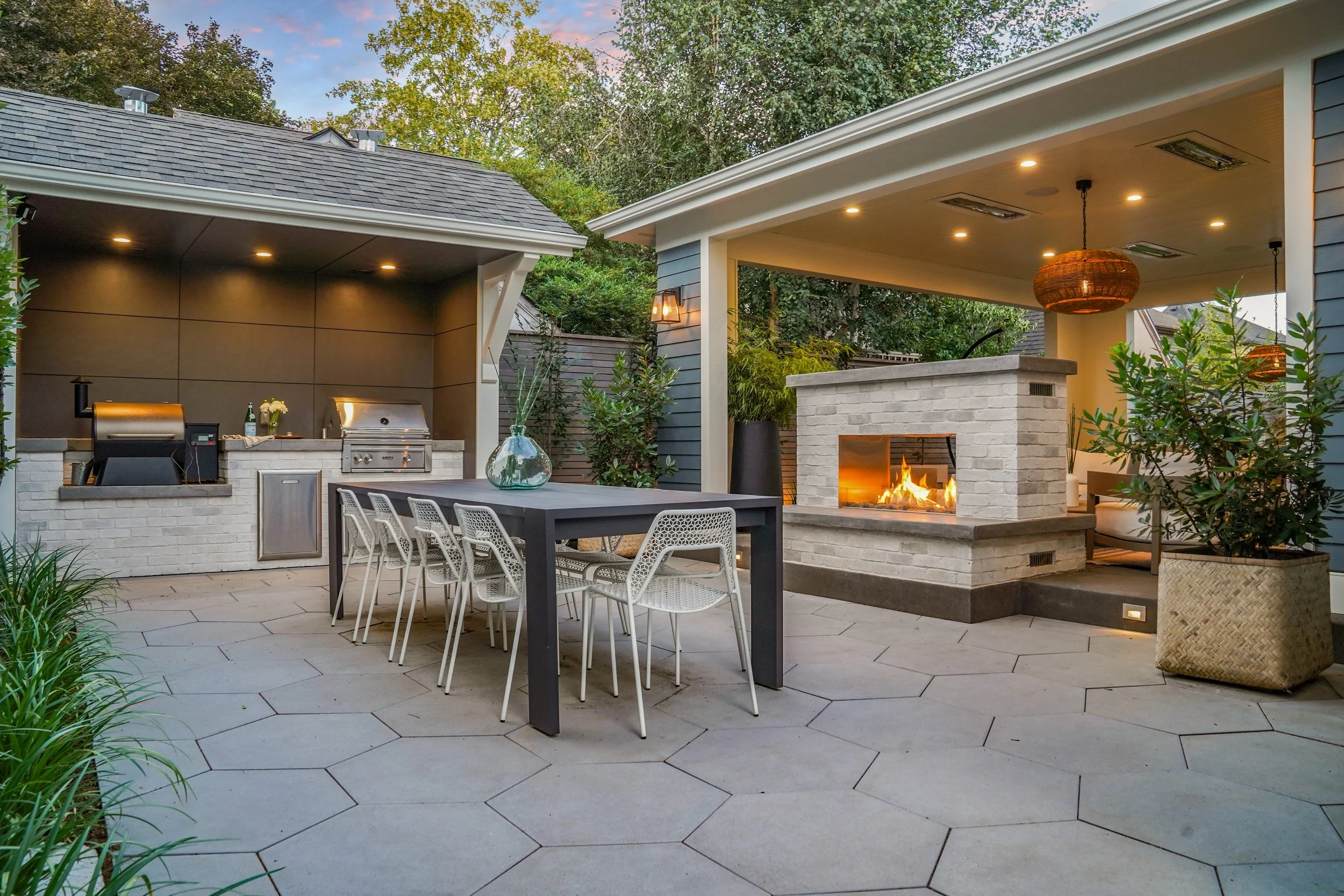 Modern outdoor patio with fireplace, dining table, chairs, built-in grill, and potted plants in a landscaped backyard.