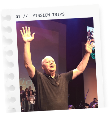 An elderly man with glasses and a black shirt raising both arms in front of a stage with a group of people, a screen, and musical instruments, under a sign that reads 'MISSION TRIPS'.