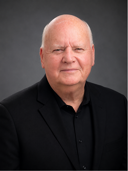 Close-up portrait of a senior man with a serious expression, wearing a black suit and black shirt, against a dark gray background.
