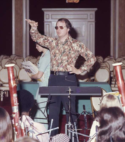 Man in sunglasses and a patterned shirt standing with one arm raised in a large room with chairs and people, possibly at a concert or event.
