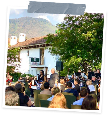 An outdoor musical performance with a conductor leading an orchestra, audience seated in front, and a building with a tiled roof in the background, surrounded by lush green trees and mountains.