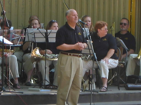 A man standing and speaking at a microphone in front of a seated band during a performance.