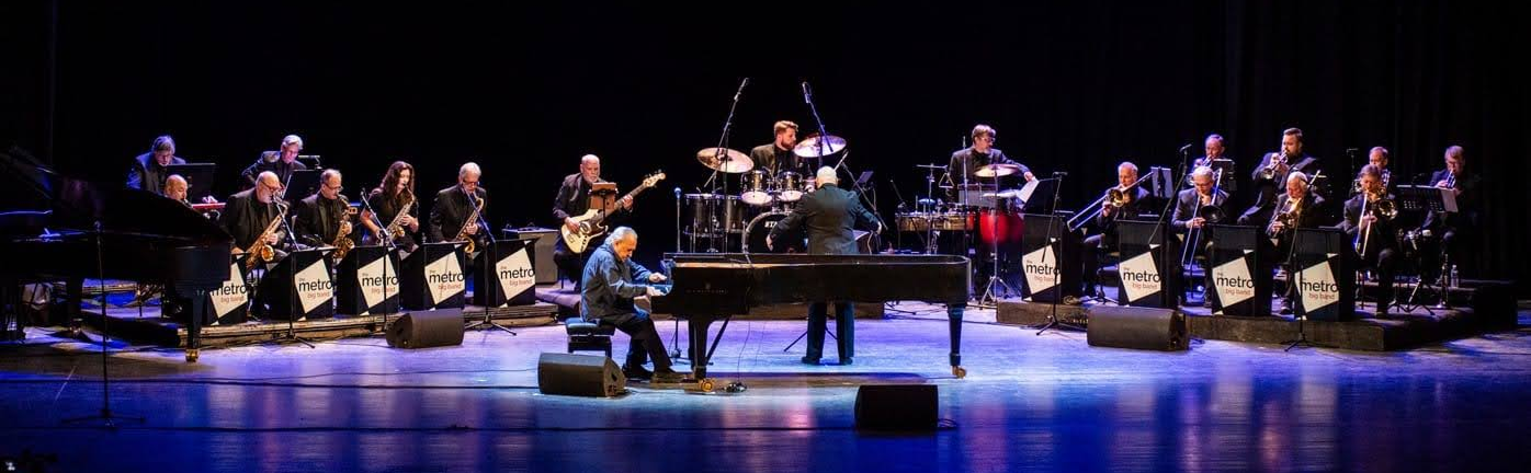 Orchestra performing on stage with a grand piano, drums, and wind instruments, with the word 'metro' on music stands.