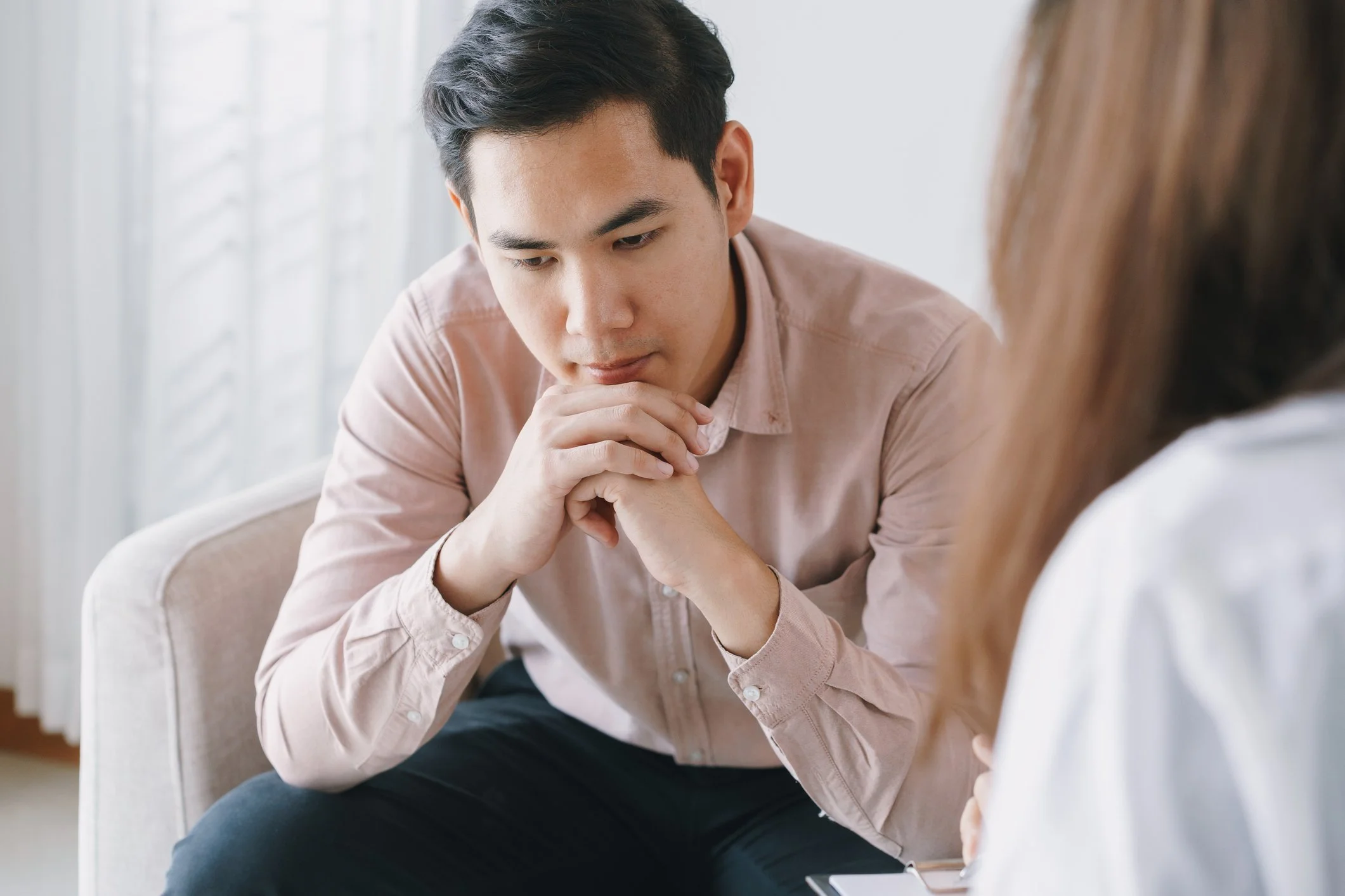 A man sitting in a counseling session witgh a woman, holding his hands together and looking down thoughtfully. 
