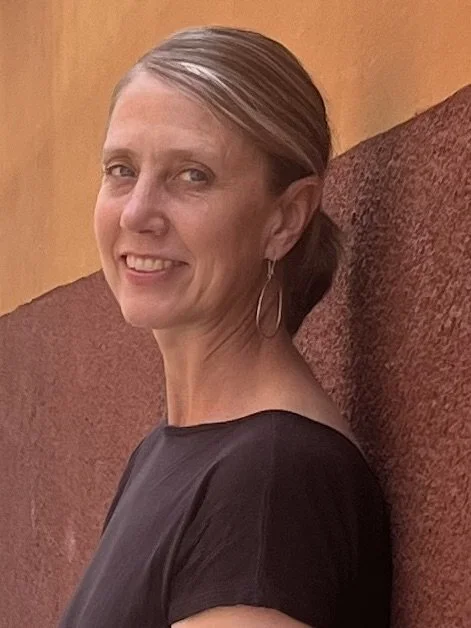 A woman with short blonde hair and hoop earrings smiling, wearing a black top, sitting against a brown textured wall.