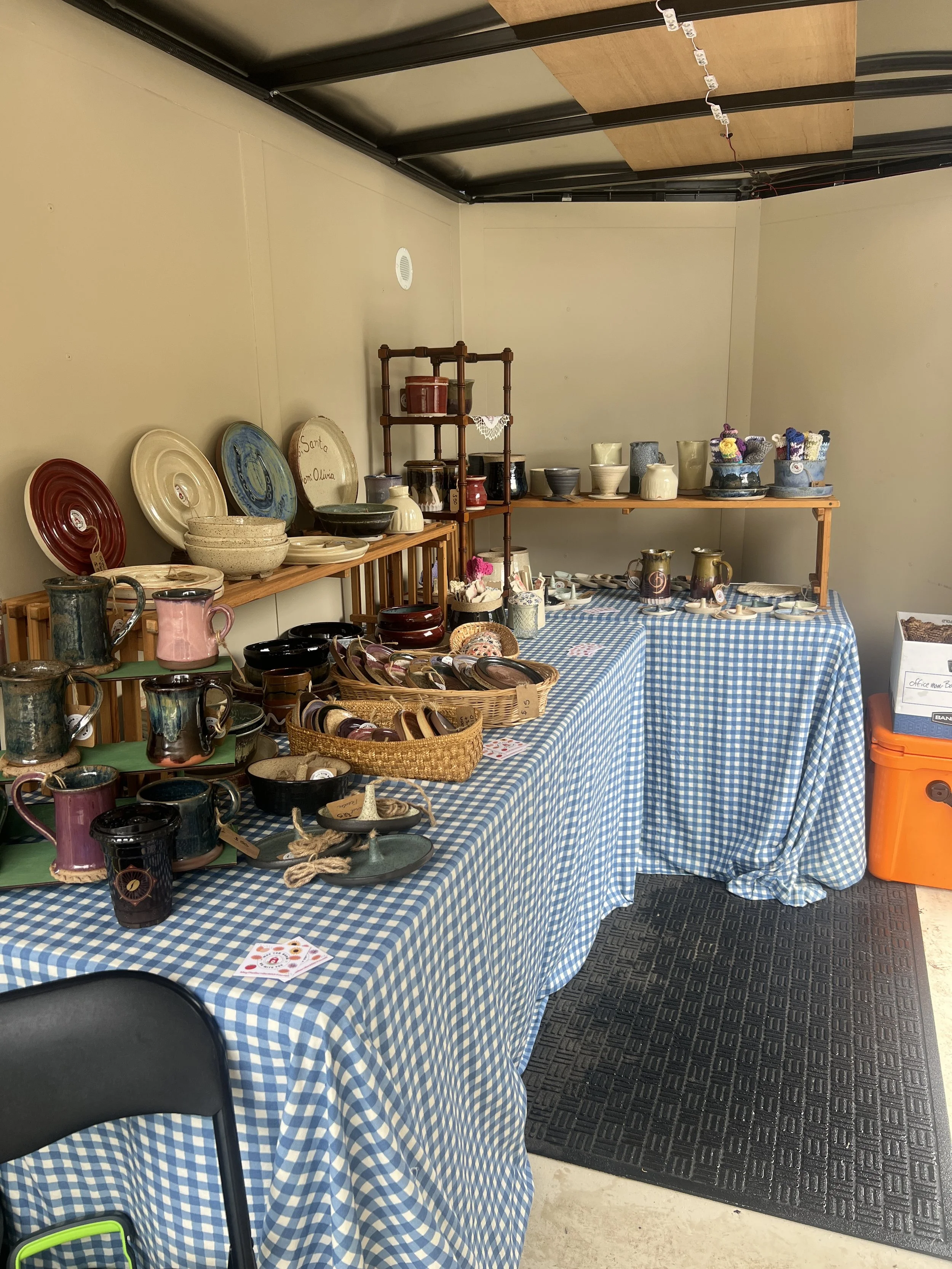 Display of various ceramic and pottery items, including plates, bowls, mugs, and vases, arranged on tables covered with blue and white checkered tablecloths at a craft or flea market.