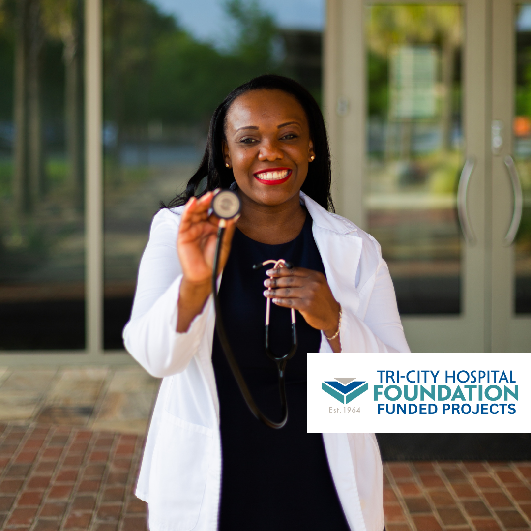 A female doctor holding a stethoscope and smiling at the camera outside a hospital entrance.