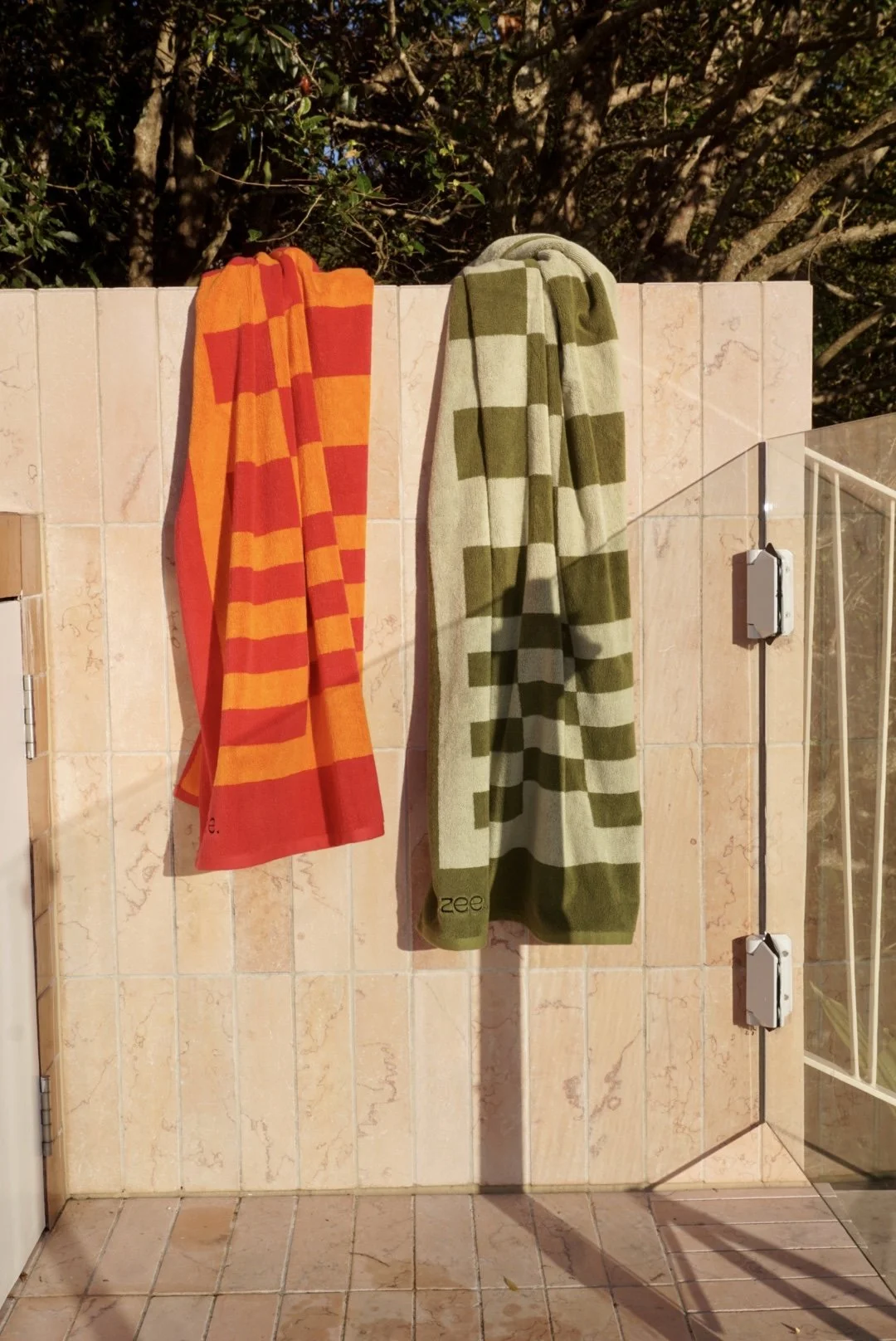 Two towels hanging on a tiled outdoor wall, one red and orange checkered towel on the left, one green and beige checkered towel on the right, with trees in the background.