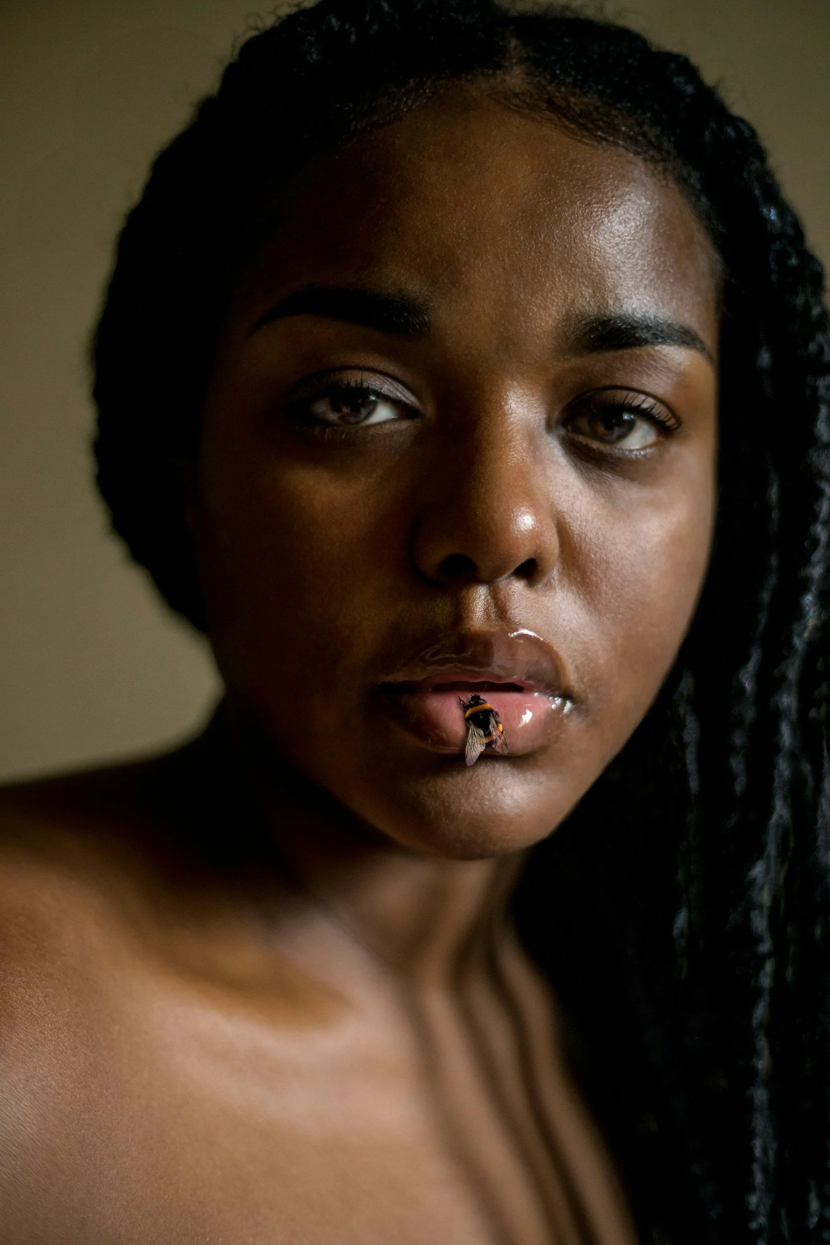 A close-up of a woman with dark curly hair, dark skin, and expressive eyes. There is a bee on her lower lip.