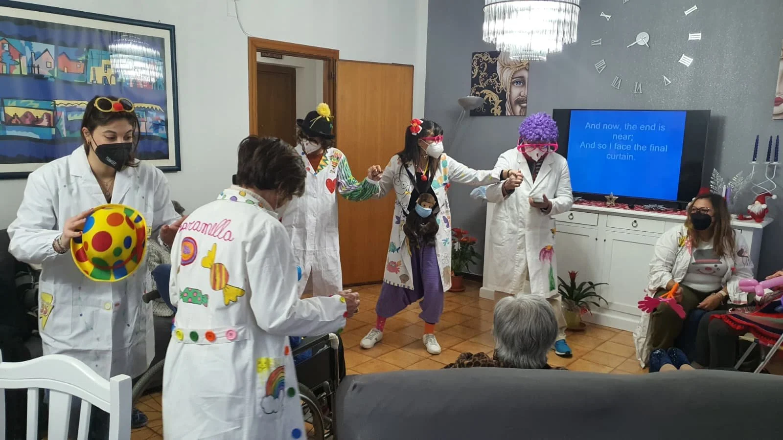 Personaggi vestiti da clown e infermieri che cantano e ballano durante una festa in una stanza decorata con luci natalizie, piante e un grande orologio sulla parete. Un schermo tv visualizza il testo di una canzone.