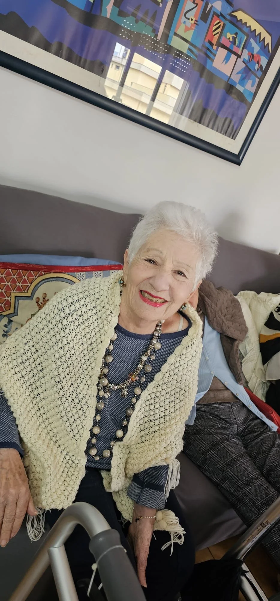 Donna anziana sorridente seduta su un divano, indossa una sciarpa color crema, una maglietta blu e una collana di perle. Dietro di lei si vede un quadro artistico astratto alle pareti.