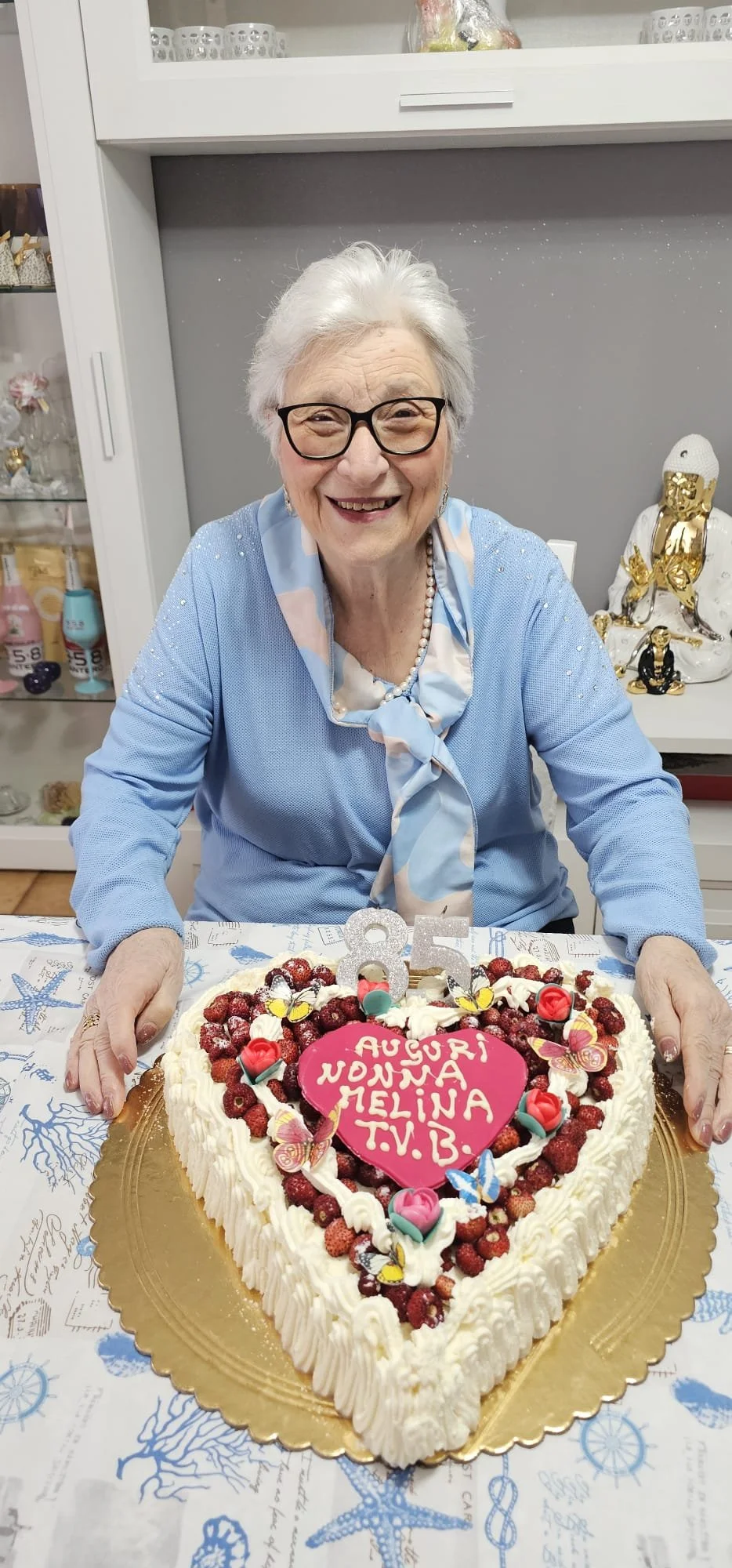Festa di compleanno con una donna anziana seduta davanti a una torta a forma di cuore decorata con frutti di bosco e decorazioni fluorescenti. La torta ha una dedica scritta in italiano e un numero 85. La donna sorride e indossa occhiali neri e una c