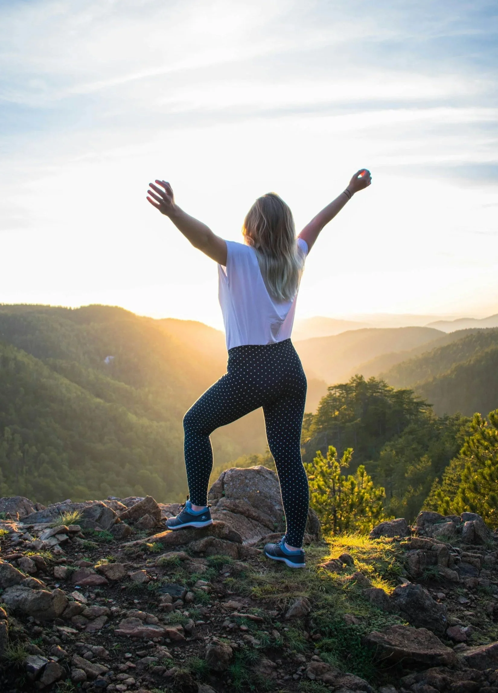 A woman standing on rocks with arms raised, facing a scenic mountain landscape during sunset.