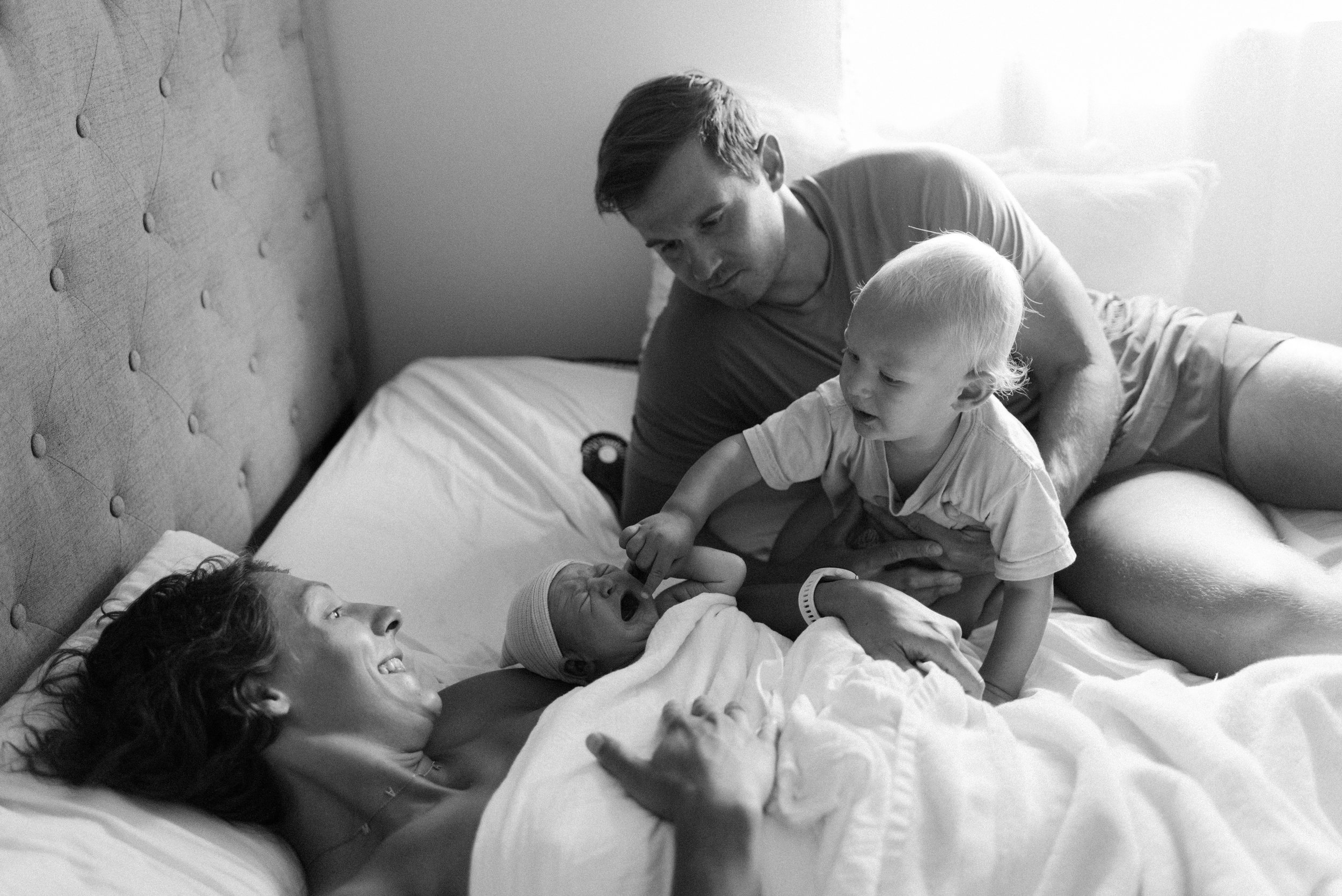 A woman and a young girl with curly hair are lying in bed, smiling and looking at a newborn baby who is being held by another child. The scene is in black and white and appears to be in a hospital or home bedroom.