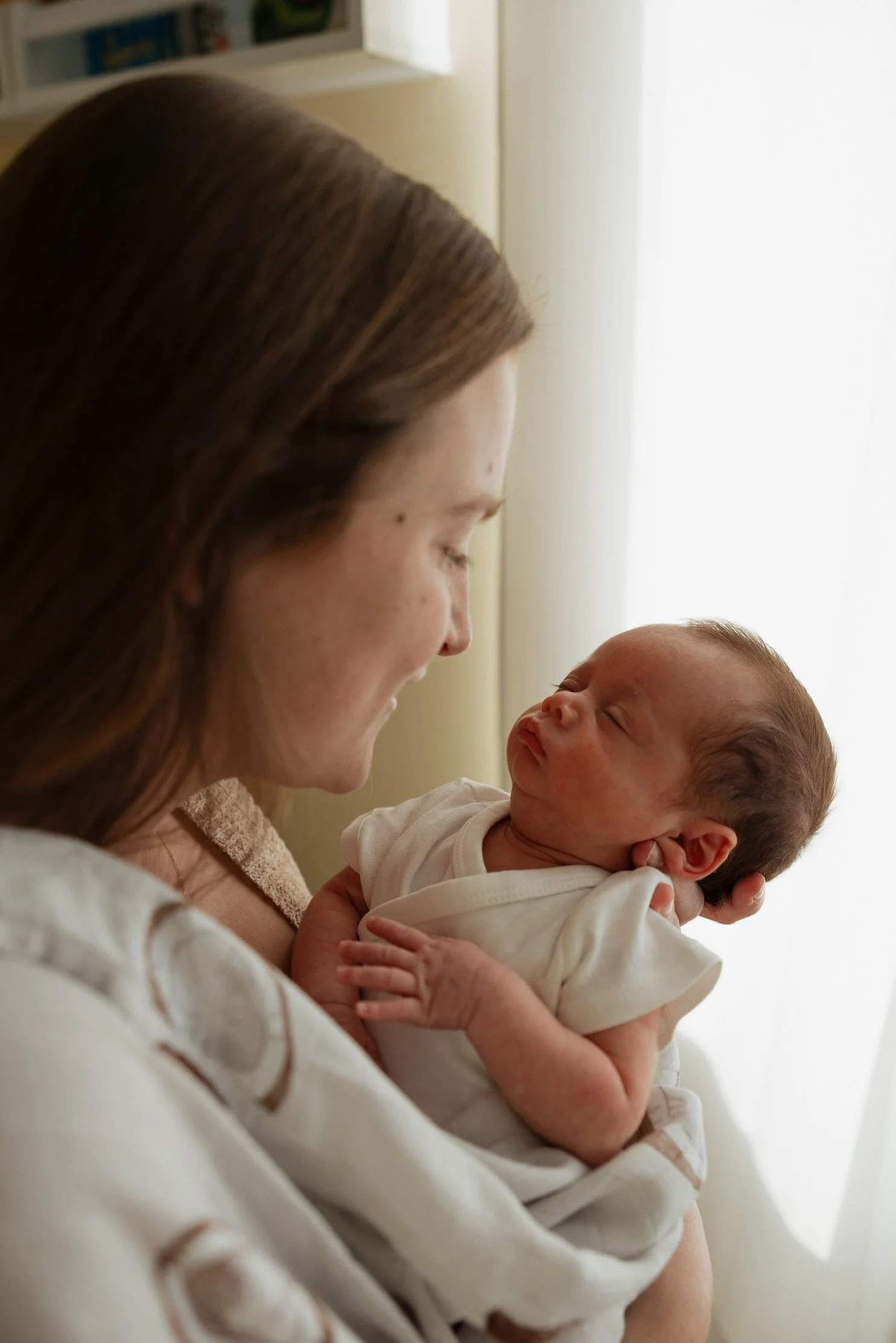 A woman with brown hair holding a newborn baby near a window with light coming through, both with closed eyes.