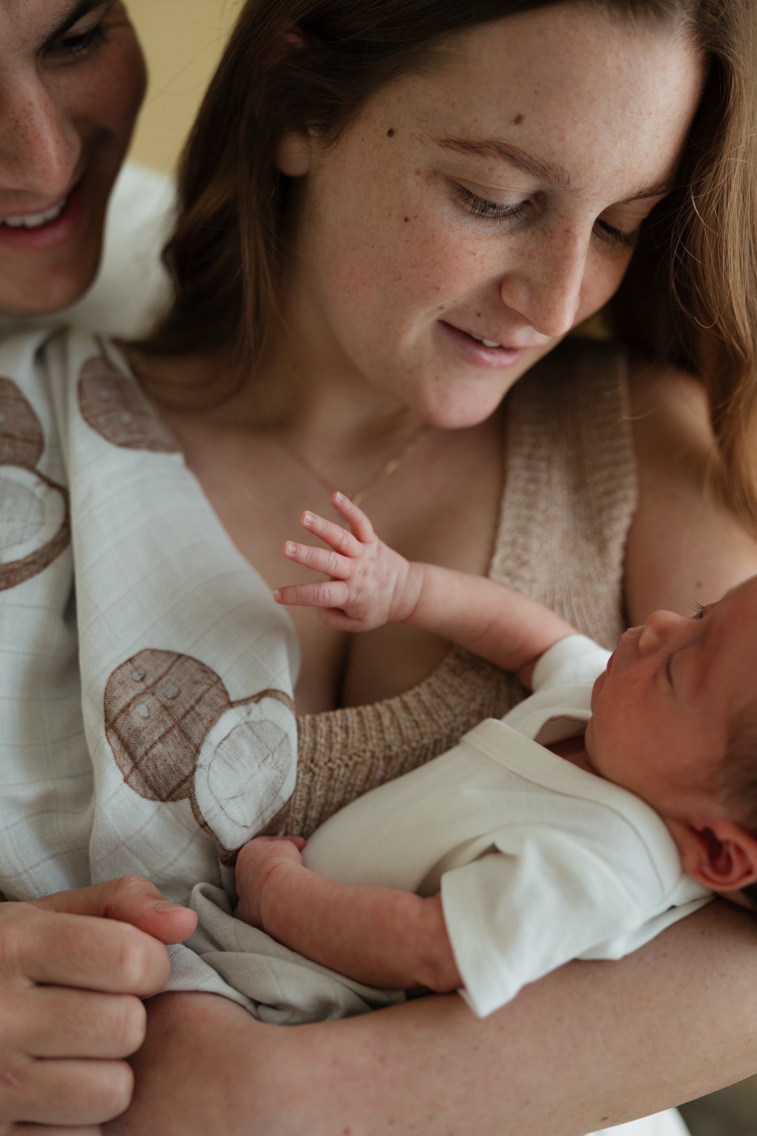 A woman with red hair and freckles holds a newborn baby, who is wearing a white outfit, while smiling softly and looking at the baby.