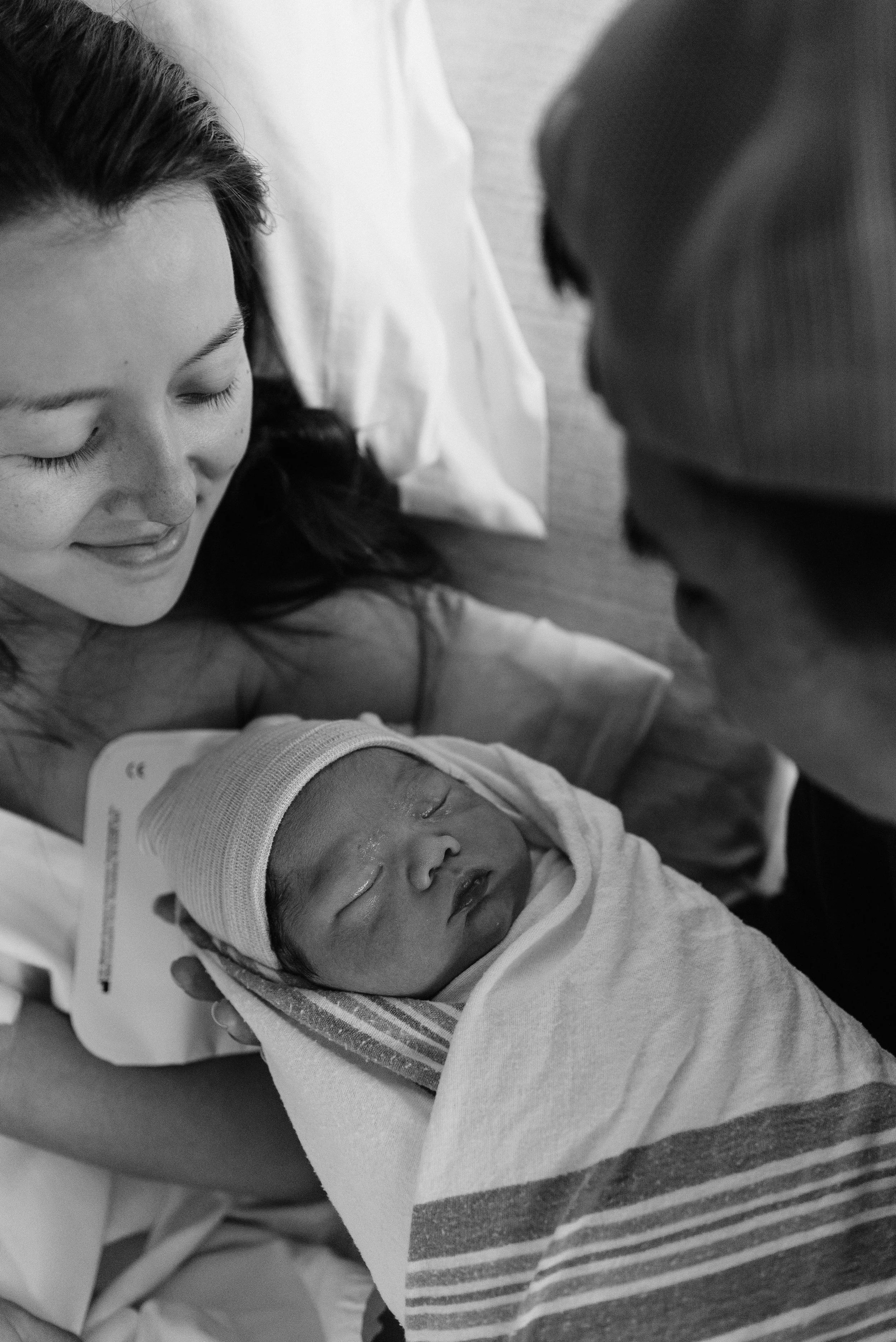 A woman in a hospital bed with a hospital wristband, holding a newborn baby wrapped in a striped blanket, with the baby wearing a nurse cap