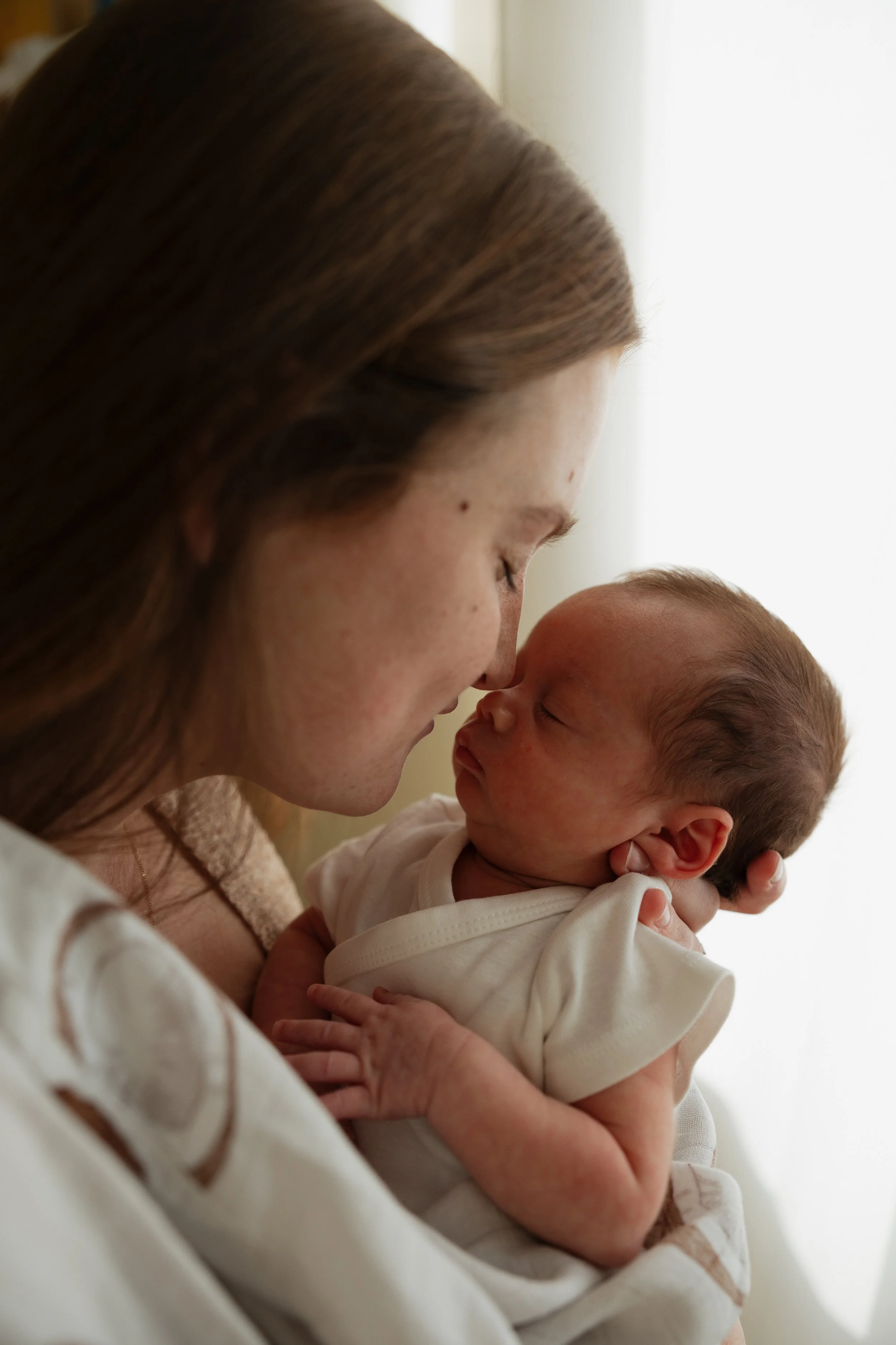 A woman holding a newborn baby close to her face, both with eyes closed, sharing an intimate moment indoors with soft lighting.