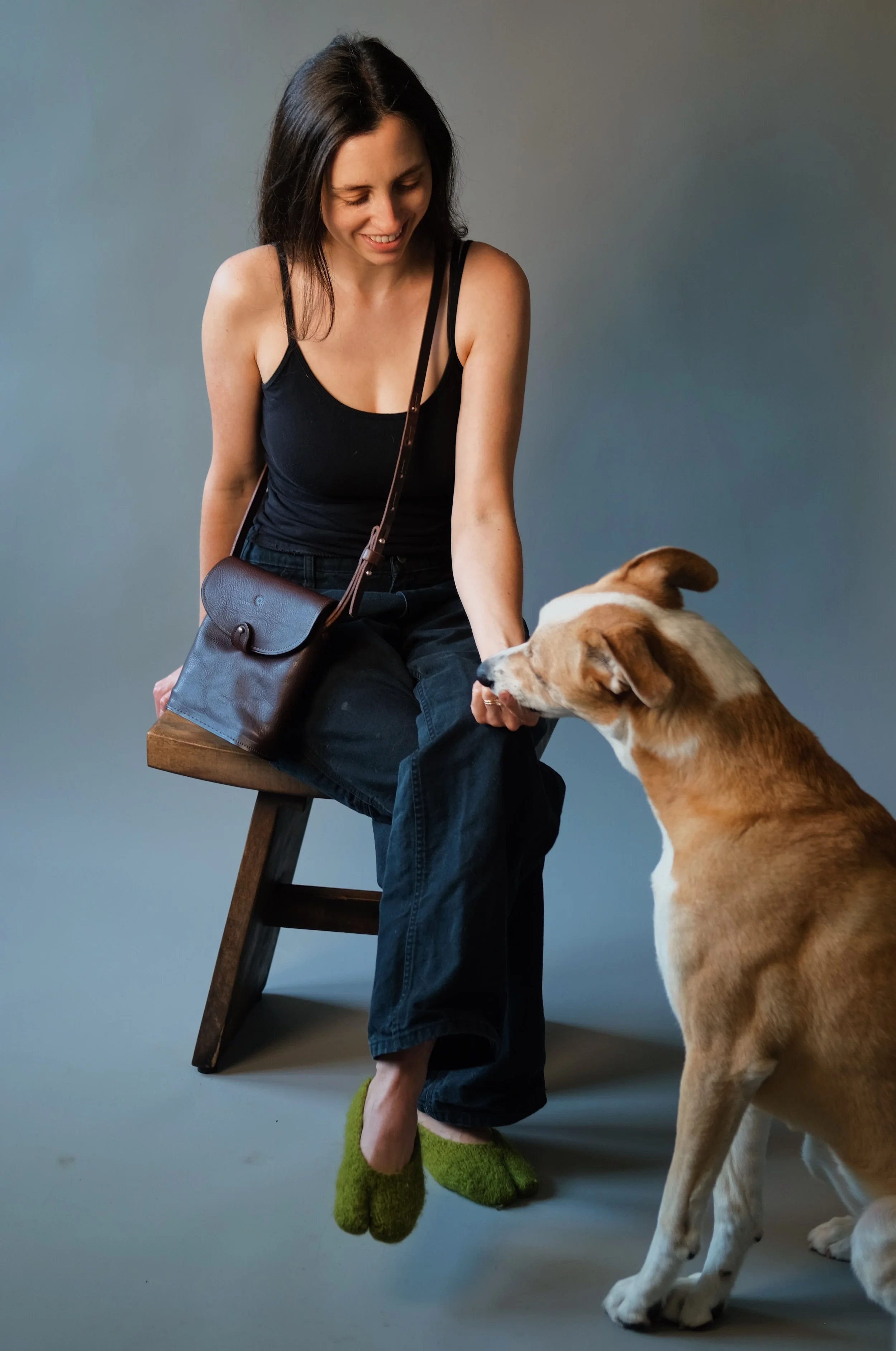A woman with dark hair sitting on a wooden stool and smiling as she pets a large dog, possibly a Labrador Retriever, against a plain gray background. She is wearing a black tank top, loose dark pants, and fuzzy green slippers, with a small brown shou