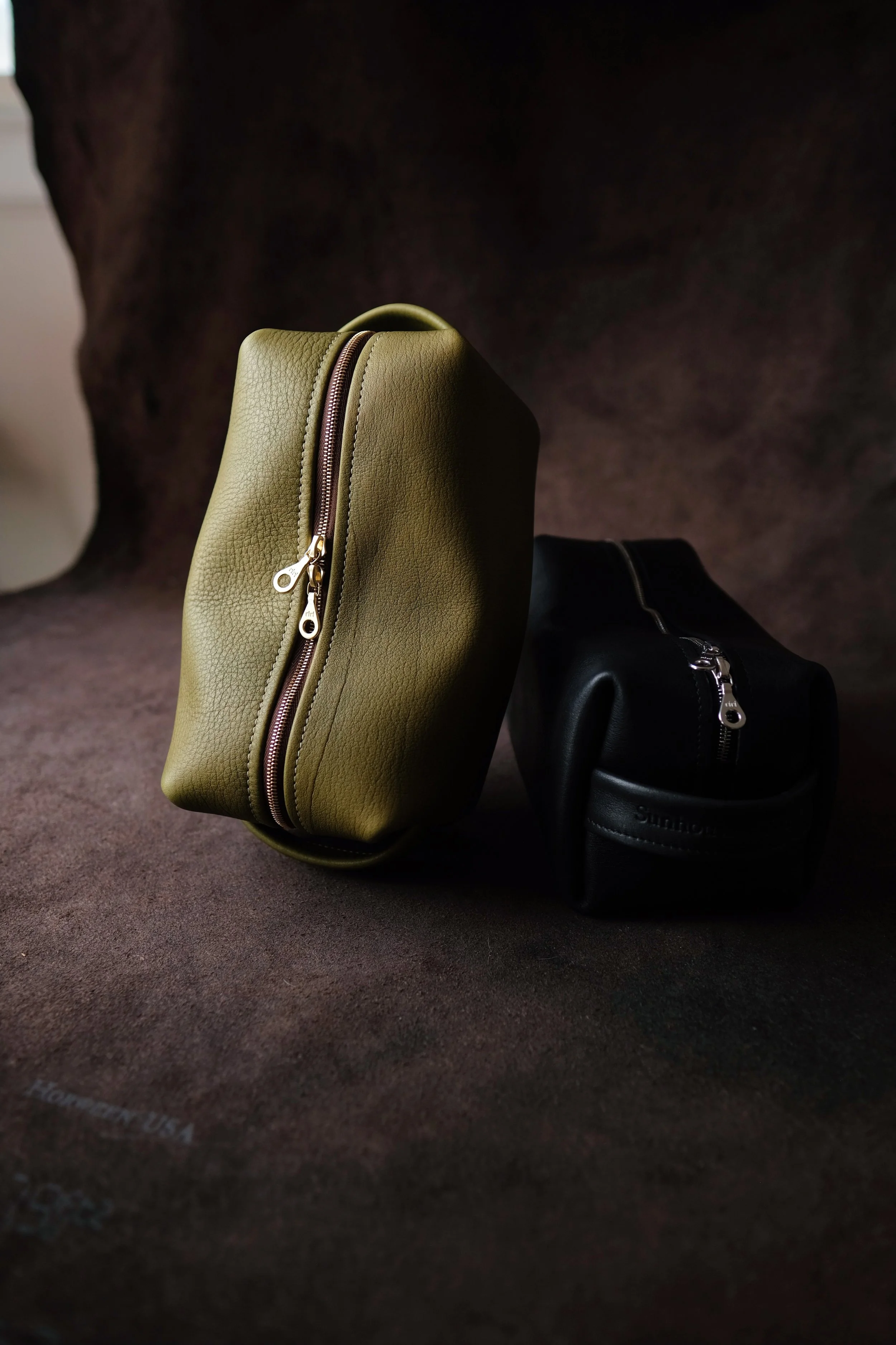 A dark brown surface displaying three small dopp kits: one green with a gold zipper, one black with a silver zipper, and the third partially visible.
