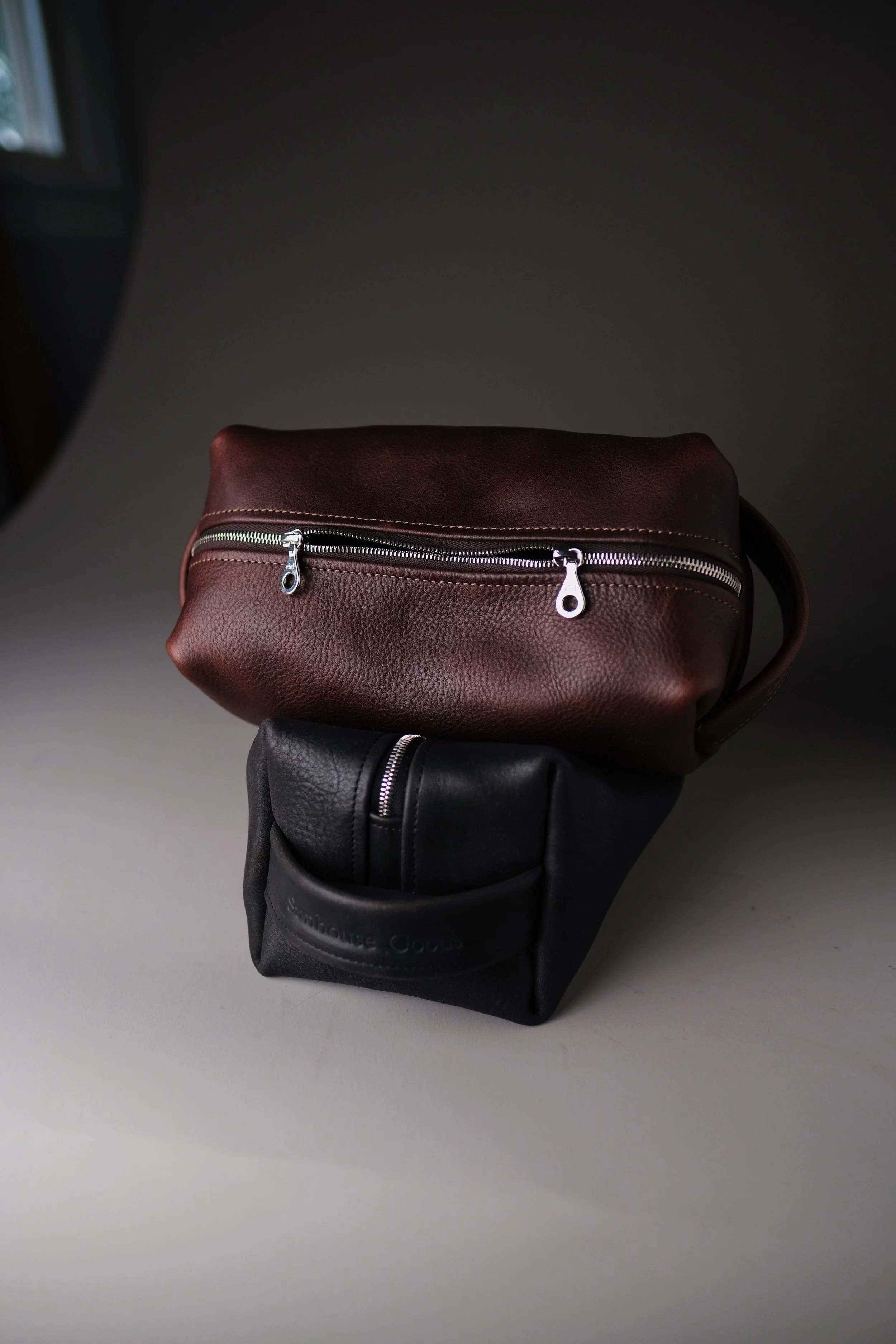 A brown leather DOPP KIT bag with a silver zipper resting on a black bag in front of a gray background.