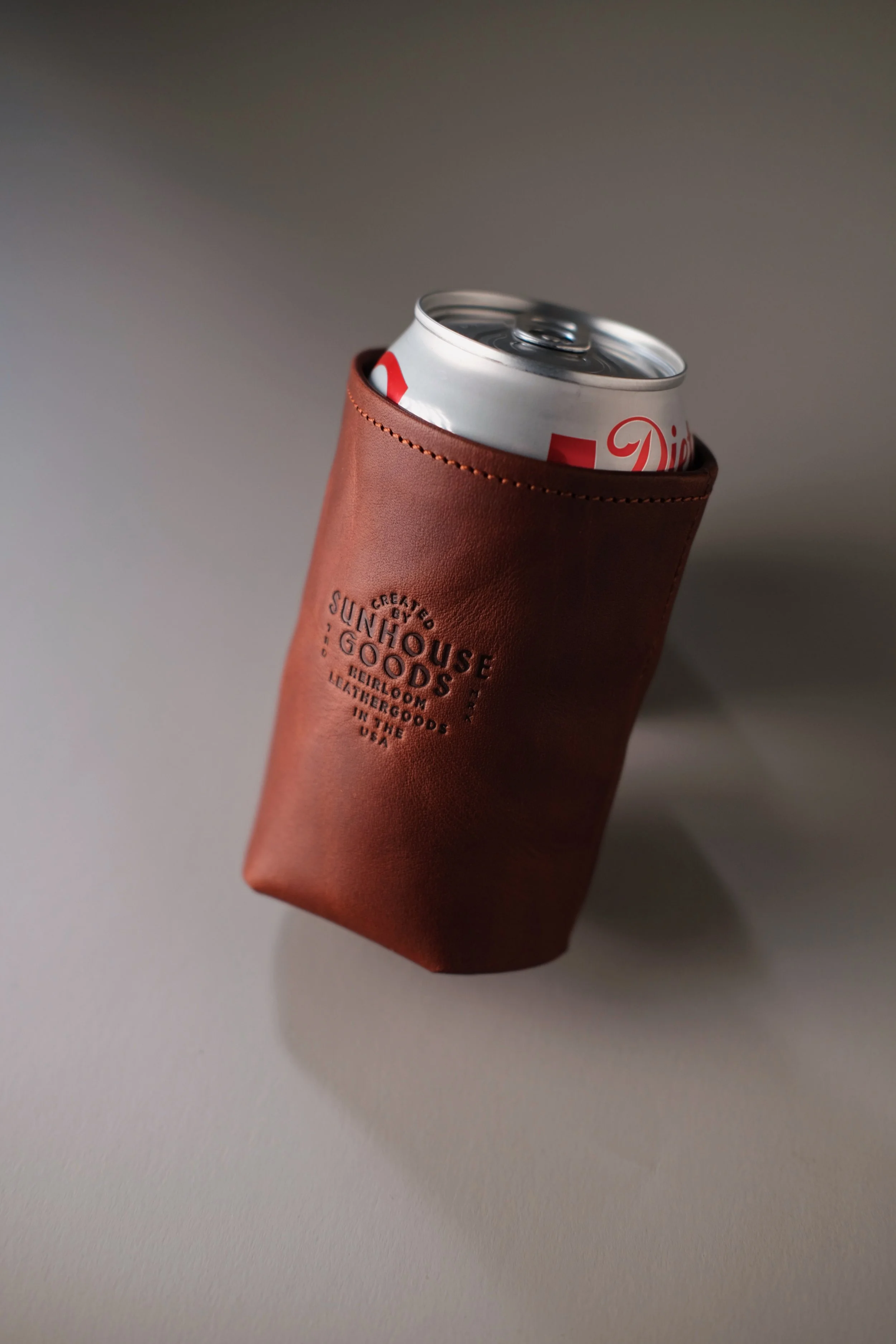 A silver soda can in a brown leather holder with embossed text that reads "CREATED BY SUNHOUSE GOODS, HEIRLOOM LEATHER GOODS IN THE USA."