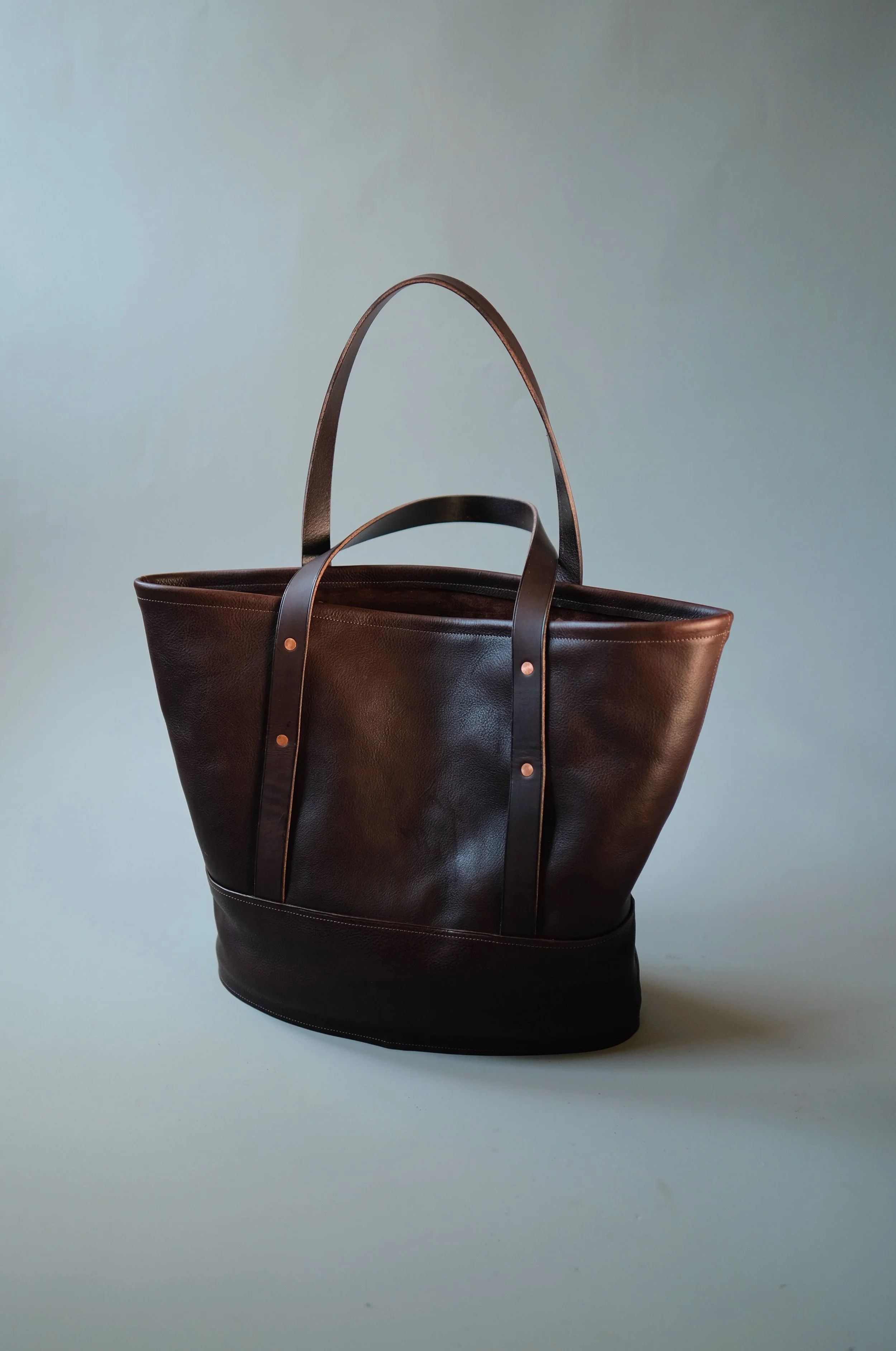 A dark brown leather tote bag with two handles, set against a simple light background.
