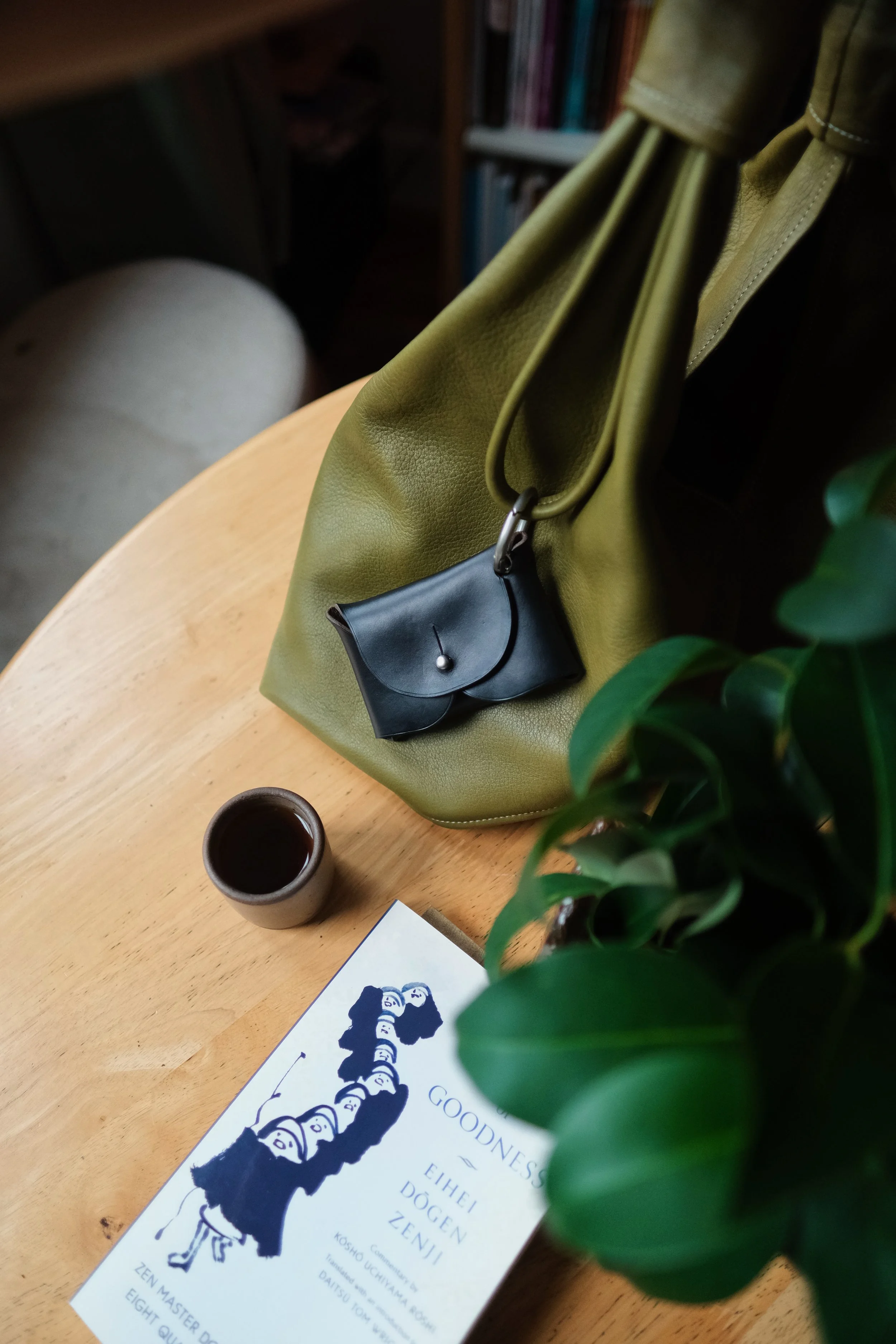 A wooden table holds a white coffee mug filled with coffee, a green tote bag with a black pouch attached, a potted plant with green leaves, and a book or brochure with a graphic of a column of faces and text in black and blue ink. In the background, there is a bookshelf filled with books and a beige or light brown upholstered chair.