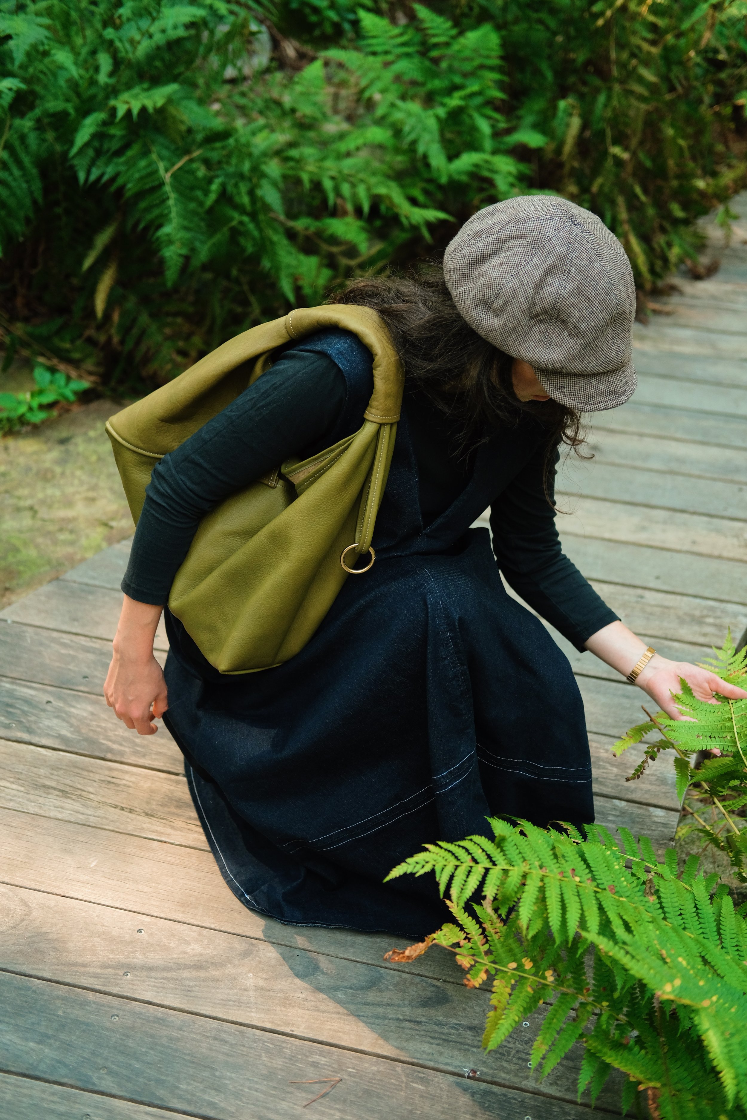 A person dressed in black with a tan bag and a gray hat, crouching on a wooden pathway in a lush, green forest, surrounded by ferns.