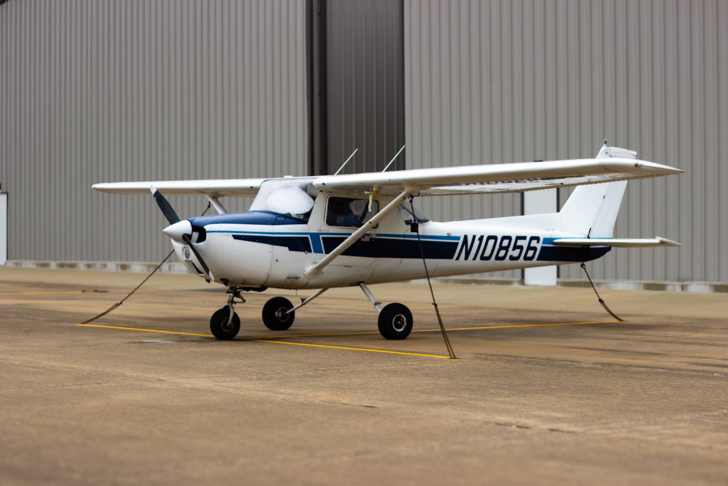 Small white and blue fixed-wing aircraft parked on an airport tarmac with a large industrial building in the background, secured with chains around its landing gear.