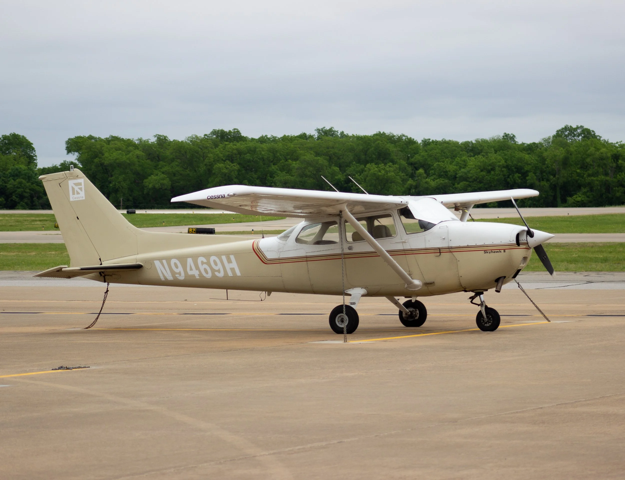 Small white light aircraft with the registration N9469H parked on tarmac, with a backdrop of trees and cloudy sky.