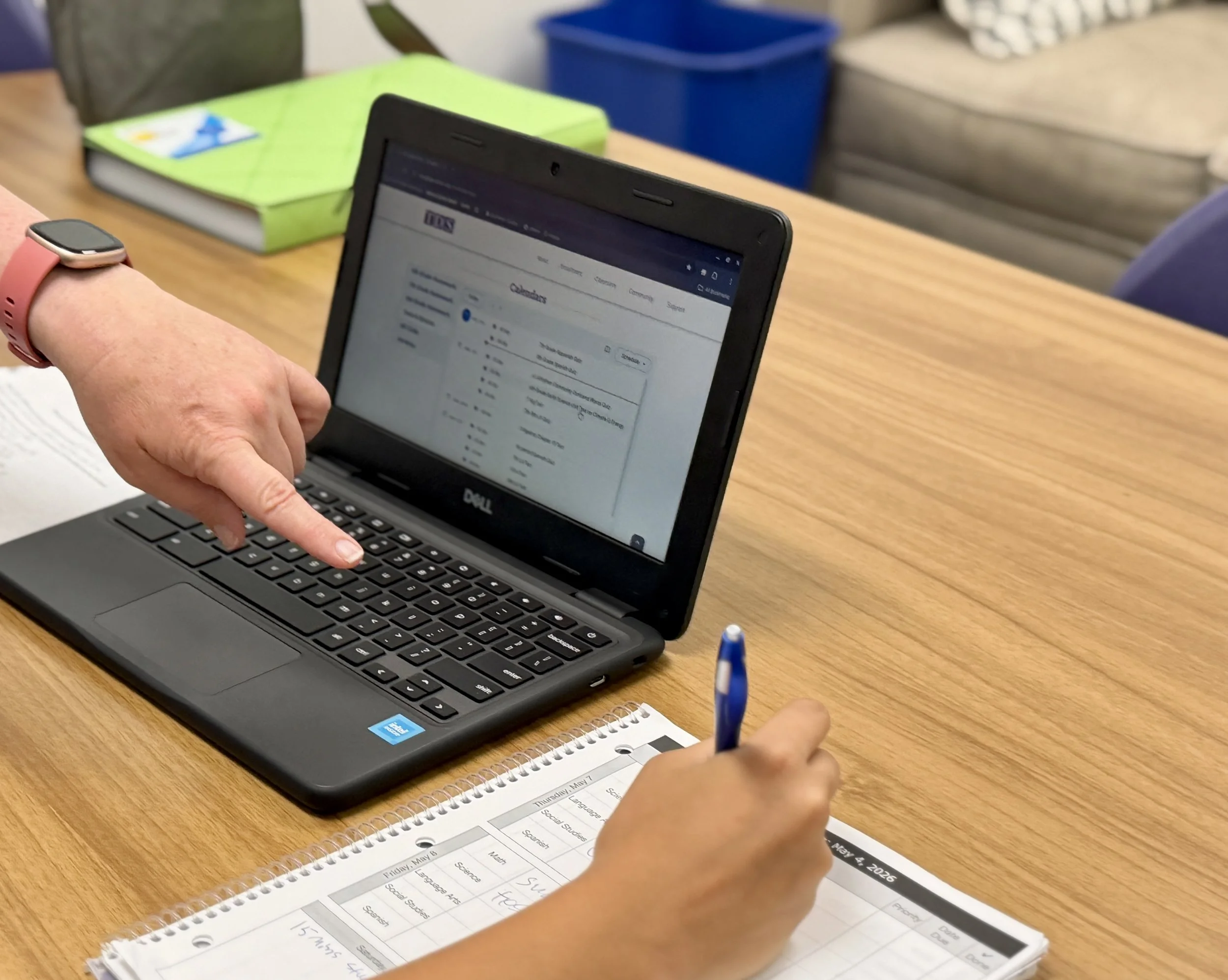 Close-up of a person's hand pointing at a laptop screen displaying a calendar; on the desk, there is a spiral planner with handwritten notes, a blue pen, and a smartwatch on the person's wrist. In the background, there are notebooks, a green one and a blue container.