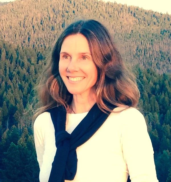 Woman smiling outdoors with forest background, wearing white top and black sweater draped over shoulders.