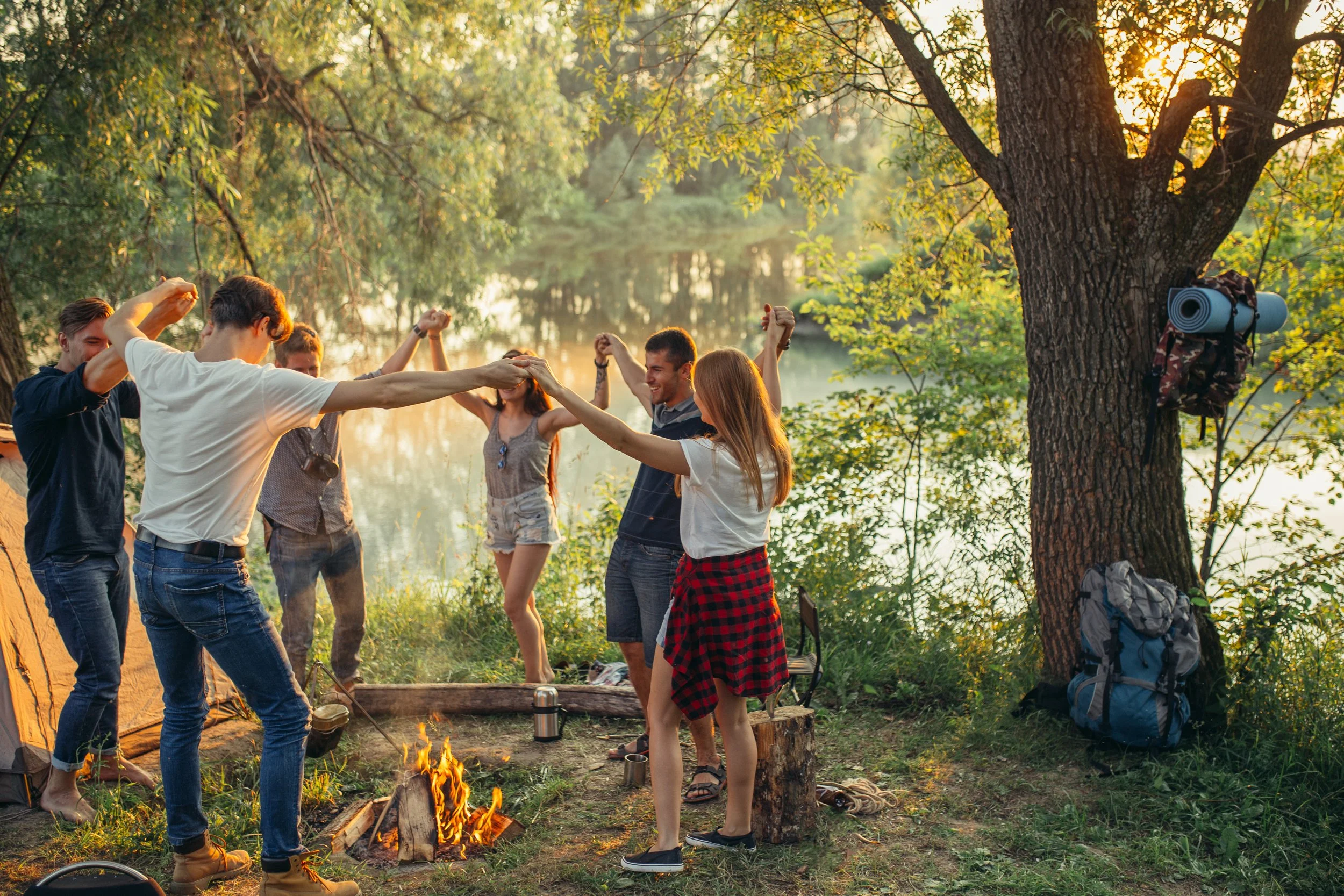 Group of people dancing around a campfire in a forest near a river or lake during sunset.