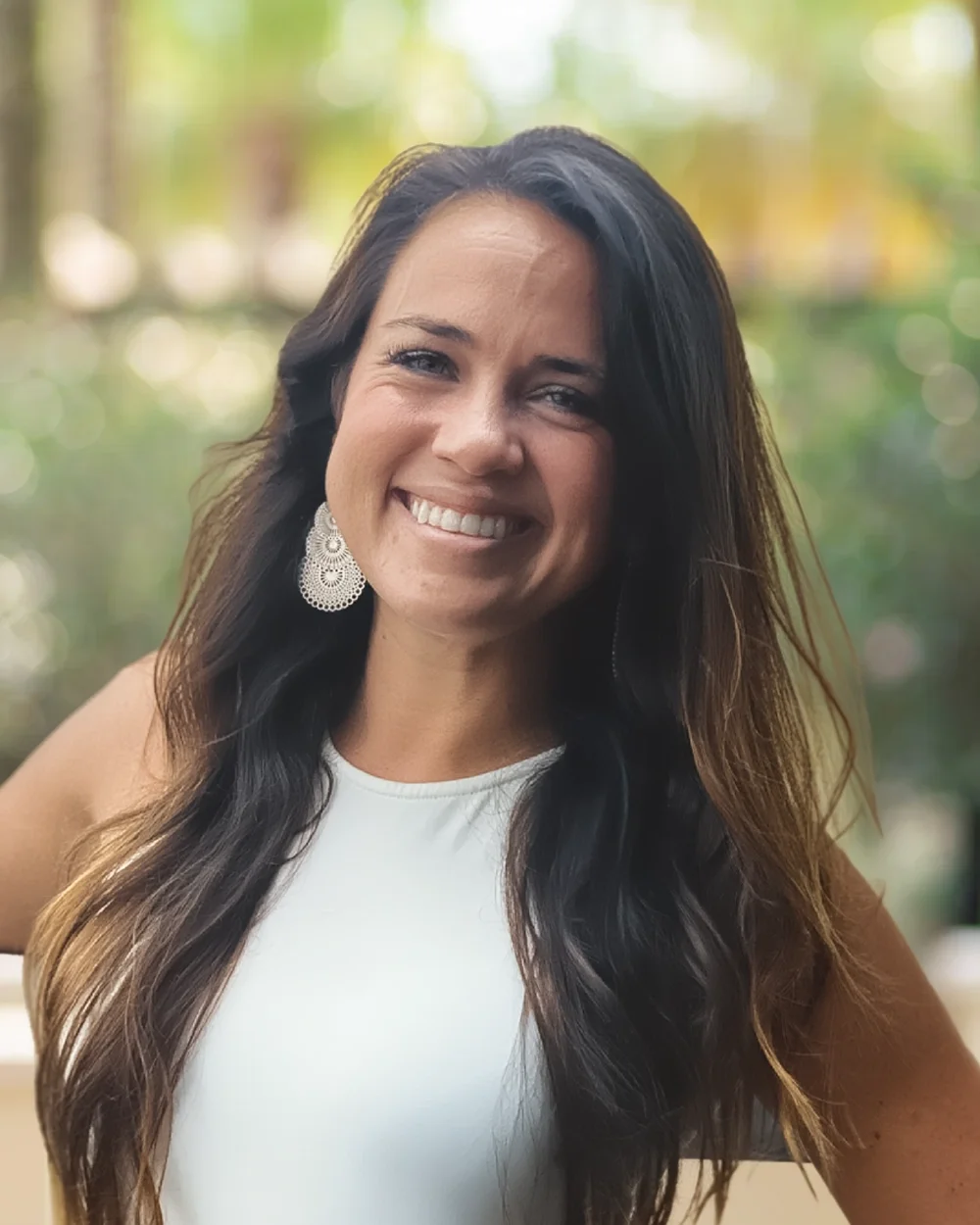 A woman with long dark hair smiling outdoors, wearing a white sleeveless top and silver earrings, with a blurred green and yellow background.