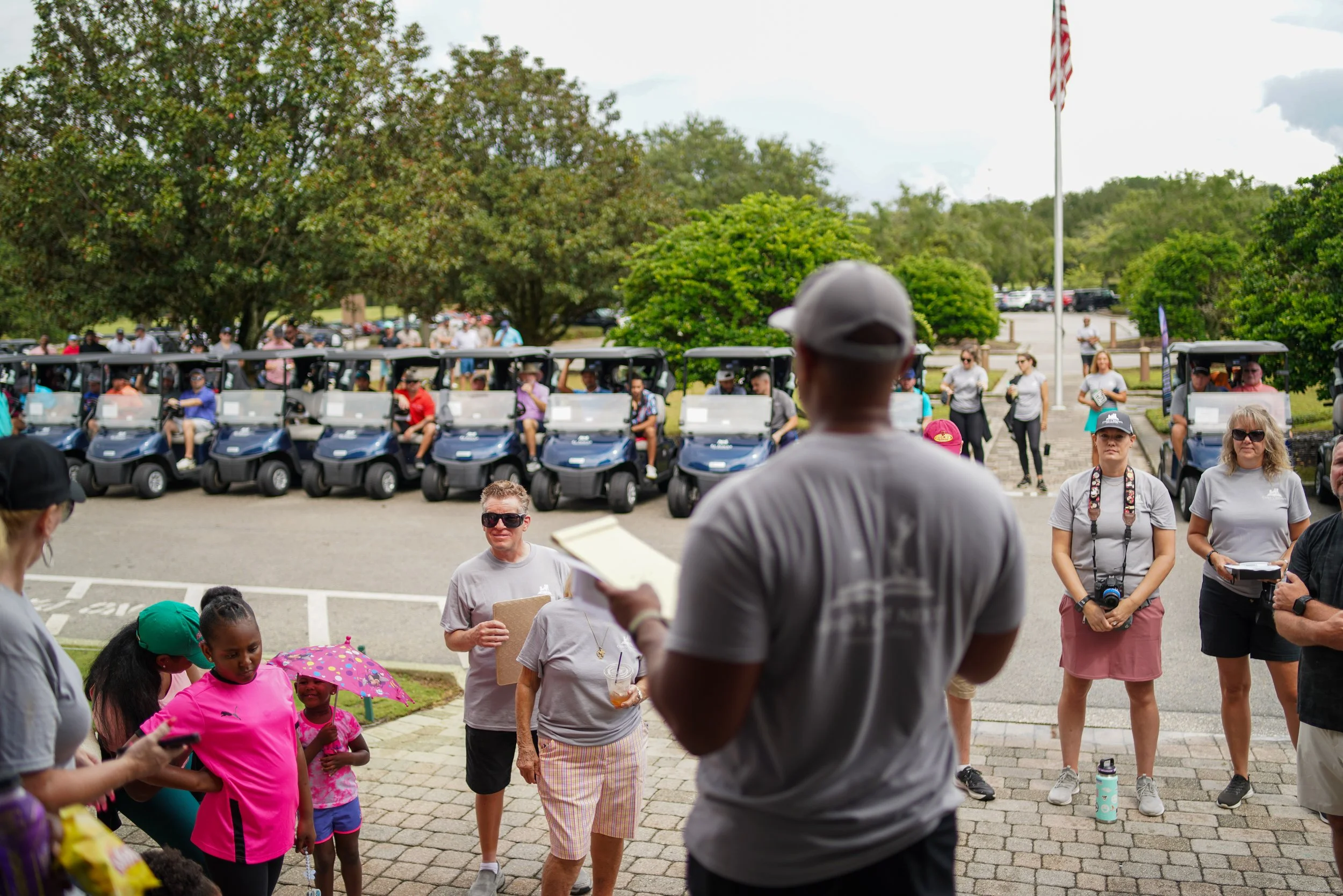 Group of people gathered outdoors near a row of golf carts, some listening to a man with a notepad, with children and adults present, on a cloudy day with trees and parking lot in the background.