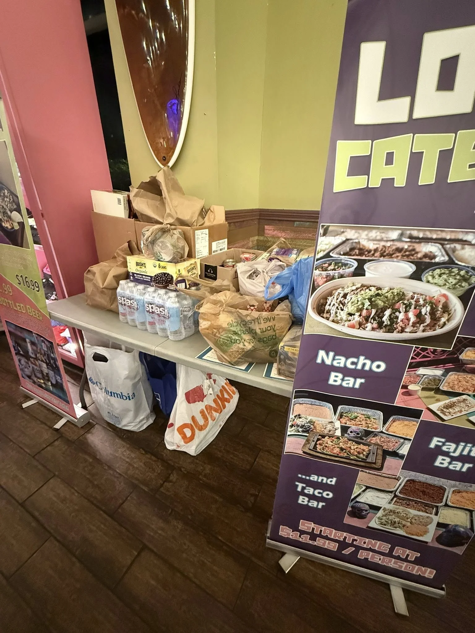 Table with grocery bags, bottled water, and snack items next to a large purple sign advertising a nacho bar, fajita bar, and taco bar at a restaurant.