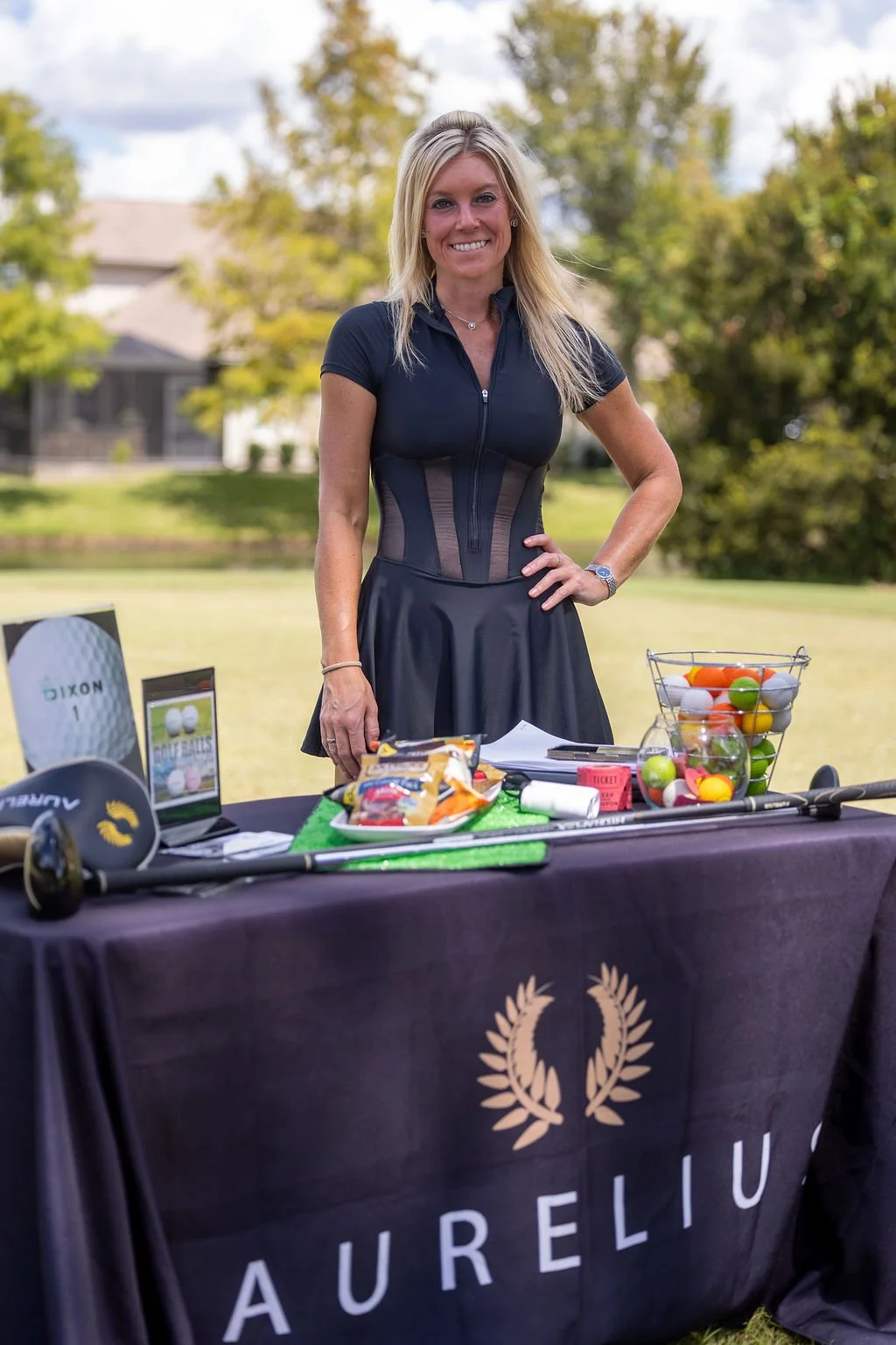 A smiling woman standing behind a table with sports equipment, golf balls, and a basket of colorful golf balls outdoors in a park with trees in the background.