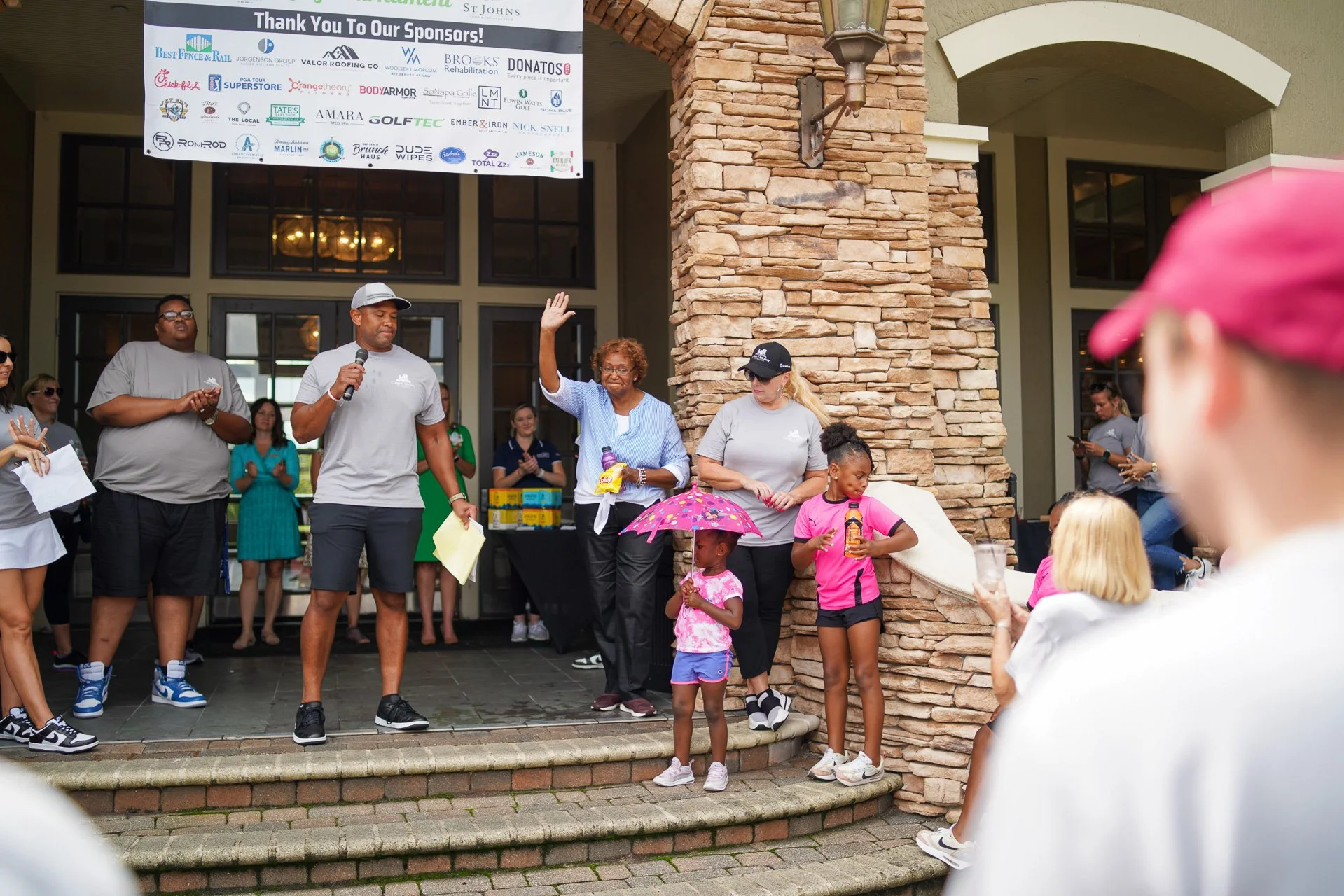Group of people on the steps of a building gathered for an event, with a woman waving and children holding umbrellas in foreground, and a large sponsor banner hanging above.