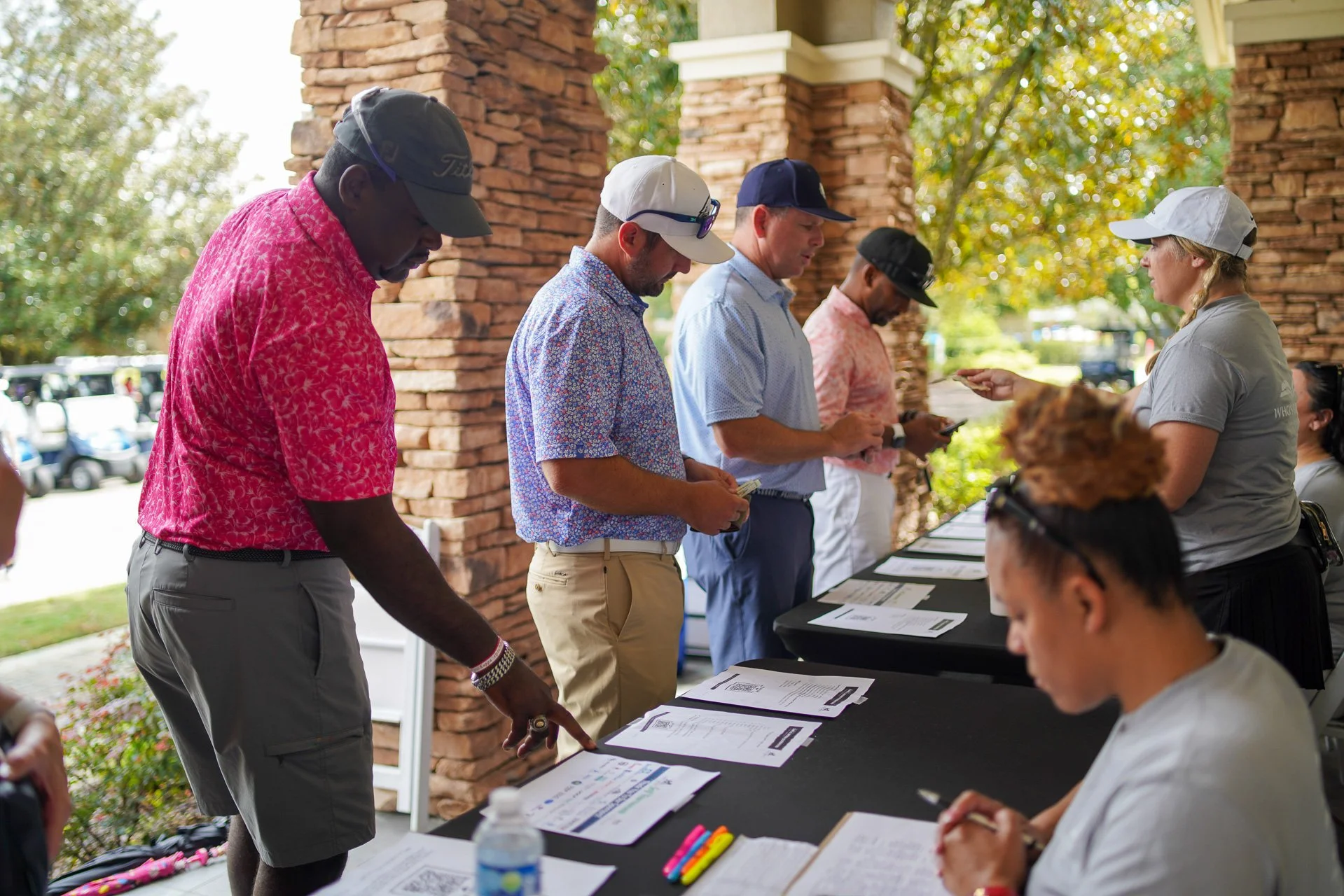 People standing in line at a registration table outside of a building with brick columns, signing in with paperwork and registration materials.