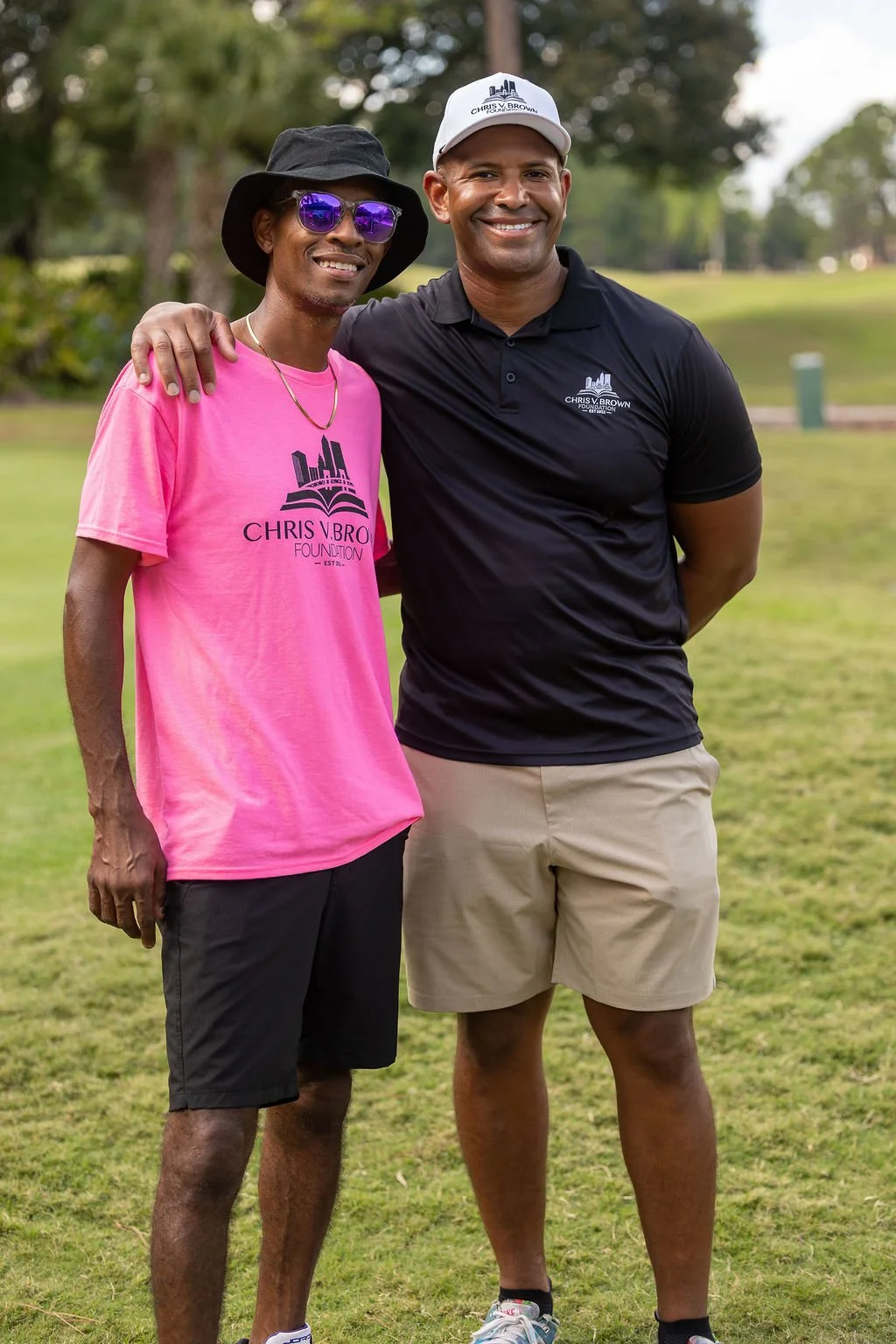 Two men standing outdoors on a grassy field, smiling at the camera. One wears a black hat, purple sunglasses, pink T-shirt with 'Chris V.Bro Foundation' logo, and black shorts. The other wears a white cap, black polo shirt with the same logo, and kha