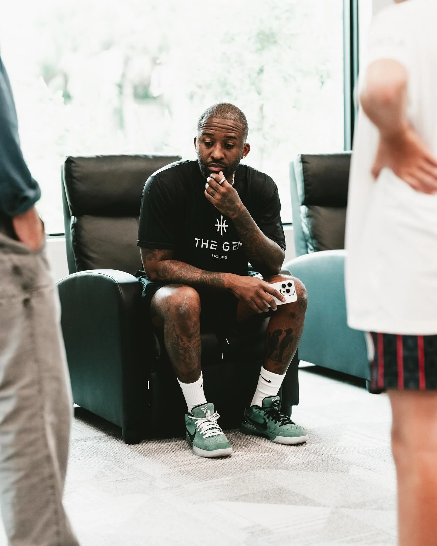 A group of people engaged in conversation at an indoor event, with a man in a black Palm Angels t-shirt in the center, surrounded by others, some in striped jerseys, and bright ceiling lights overhead.